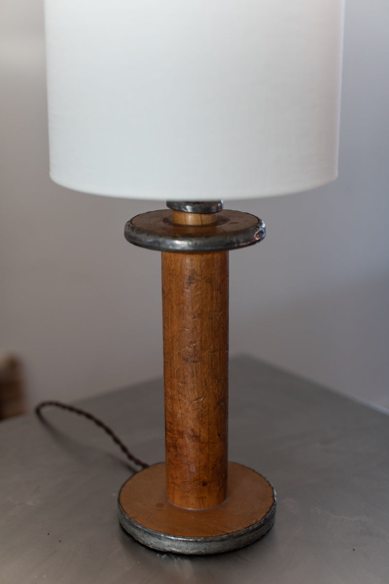 The Large Mid-Century Lamp by Out For Lunch boasts a cylindrical wooden handle base accented with steel edges. It features metal disc accents near the top and bottom, paired tastefully with a simple, white cylindrical lampshade. This elegant lamp is perfectly situated on a metallic surface.