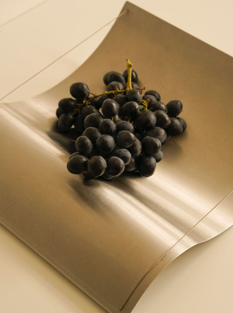 A bunch of dark grapes resting on a Les Objets Amsterdam Studio Eldink holder with a soft, cream-colored background. The lighting creates a gentle shadow around the grapes.