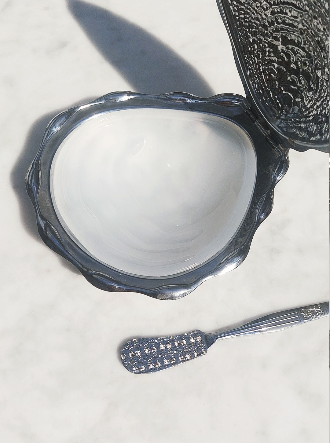 A vintage metal compact with a partially open lid revealing a white, glossy interior rests on a white marble surface. Next to the compact is the Shell Butter Dish by Les Objoies and a small, intricate metal spatula.