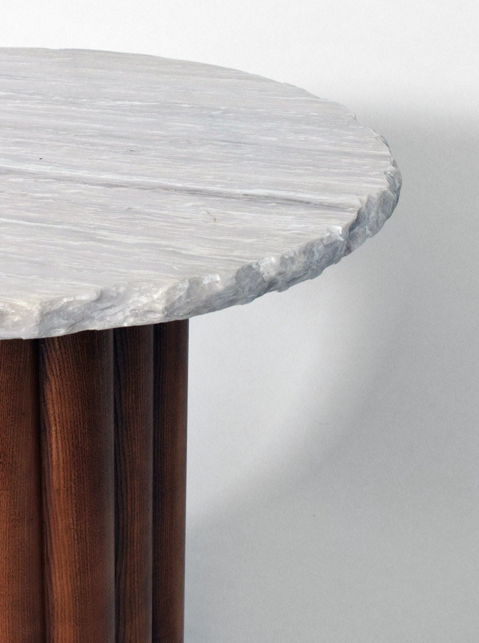 A close-up of the "Carrasqueira" table, Barracuda edition, by Barracuda Interiors with a textured, light gray stone top and an oak wooden round base made of vertical slats. The stone top has a rough, uneven edge, contrasting with the smooth, dark wooden sections. The background is plain and neutral.