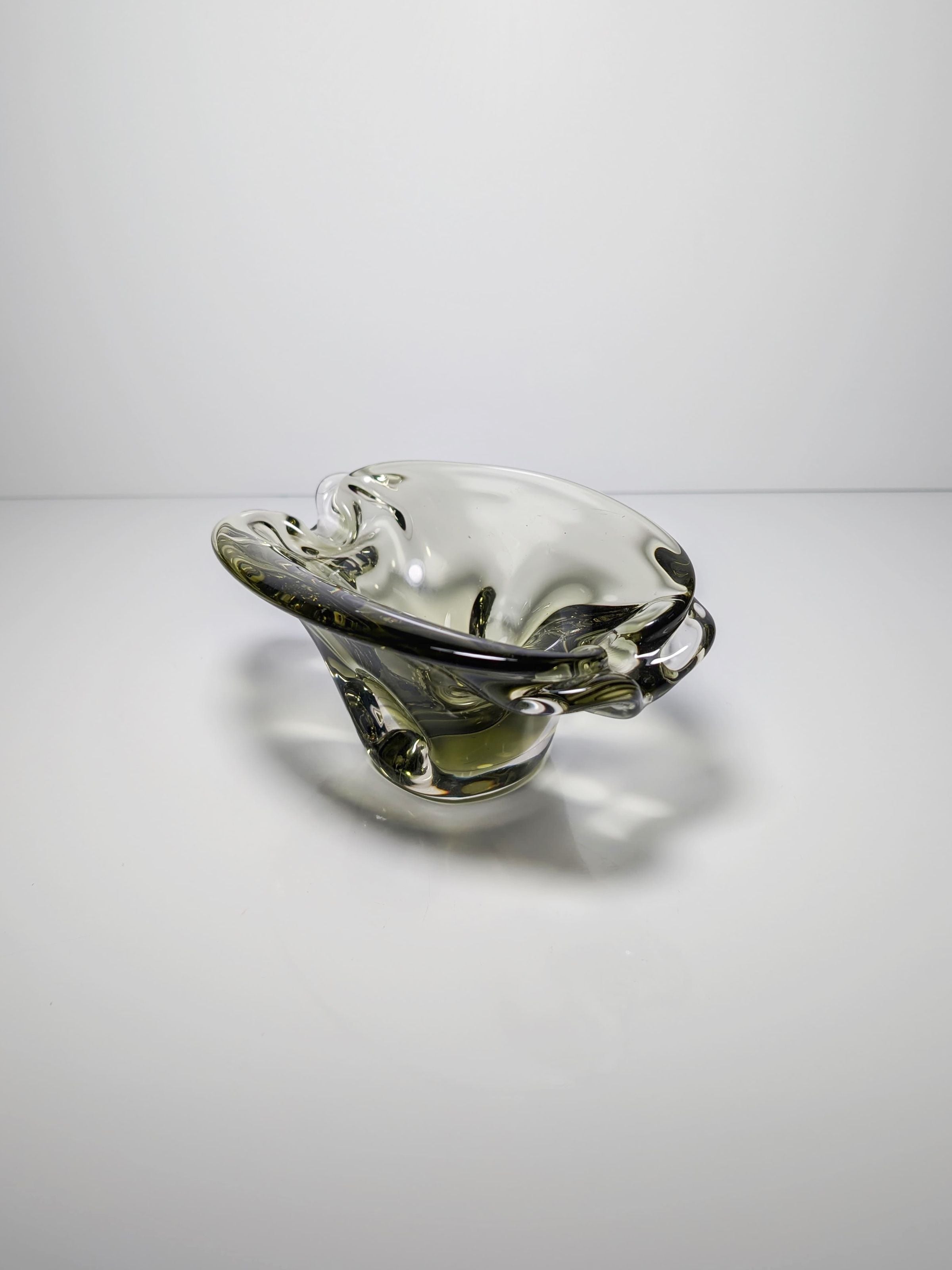 A Bohemian Smoked Glass Ashtray by Jan Beránek from the 1960s, sold by Dodo Vintage, rests on a reflective surface. Its abstract design and smooth curves are enhanced by light, creating subtle shadows and reflections.