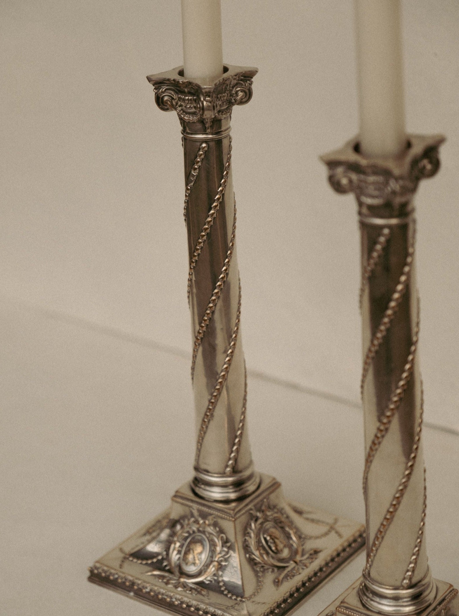 A close-up of two ornate silver candlesticks with spiral detailing and intricate designs on the bases. These Casita Amada Pair of Column Sheffield silver plated candleholders, attributed to John Winter & Co, hold slim, unlit white candles and are set against a light, neutral background.