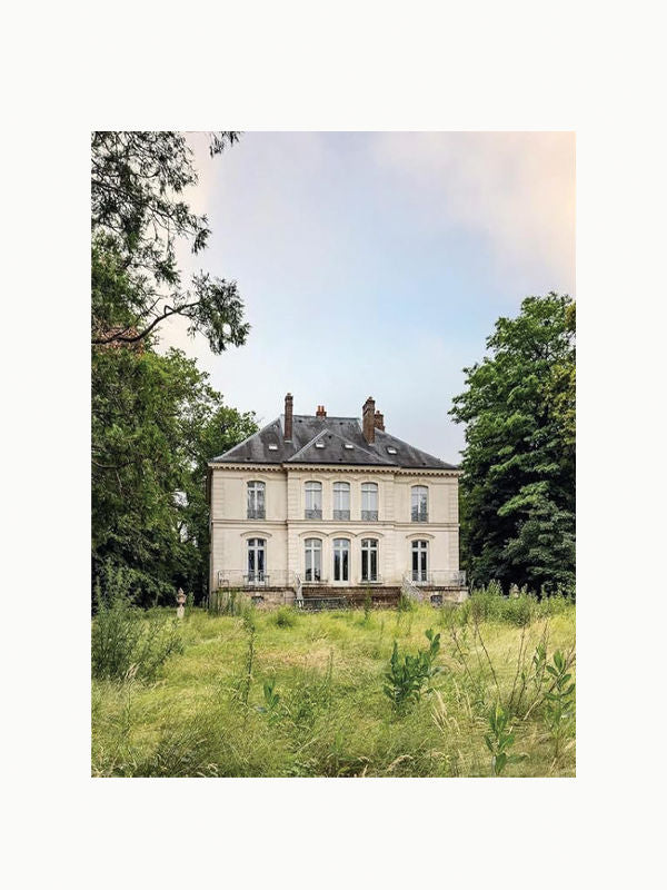 A stately, two-story mansion with elegant French architecture sits amidst a lush, overgrown field. Reminiscent of Maison Plage's Karl Lagerfeld: A Life in Houses, the house features large windows, a mansard roof, and is flanked by tall, leafy trees. The sky above is clear with a hint of pale hues.