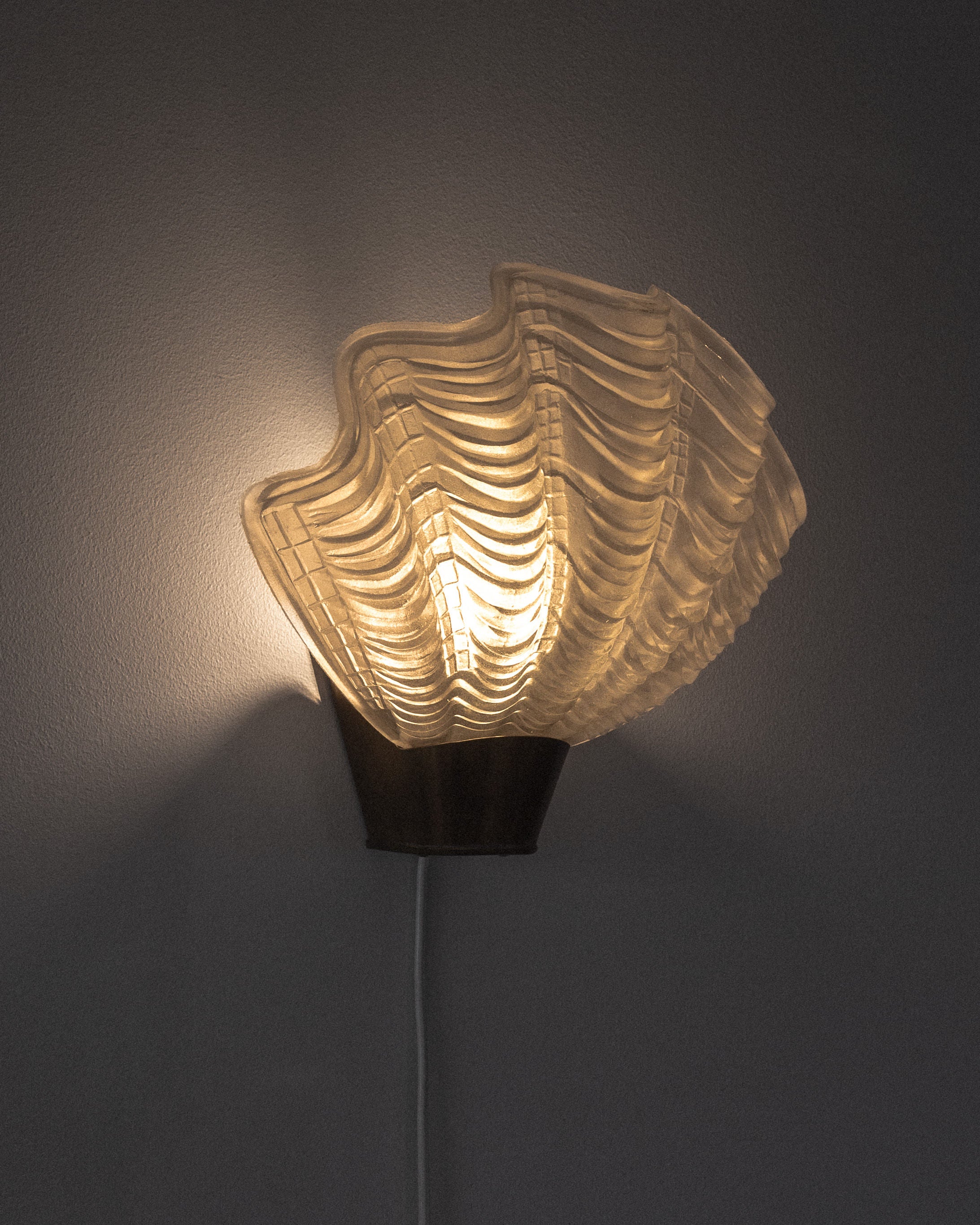 The Vintage "Coquille" Wall Light by Spigel, crafted in 1940s Sweden, features a pleated shell-like glass shade that casts a warm glow on the gray wall. Its Art Deco charm enhances the curved texture, creating a soft ambient effect with an elegantly hanging cord from the lamp's base.