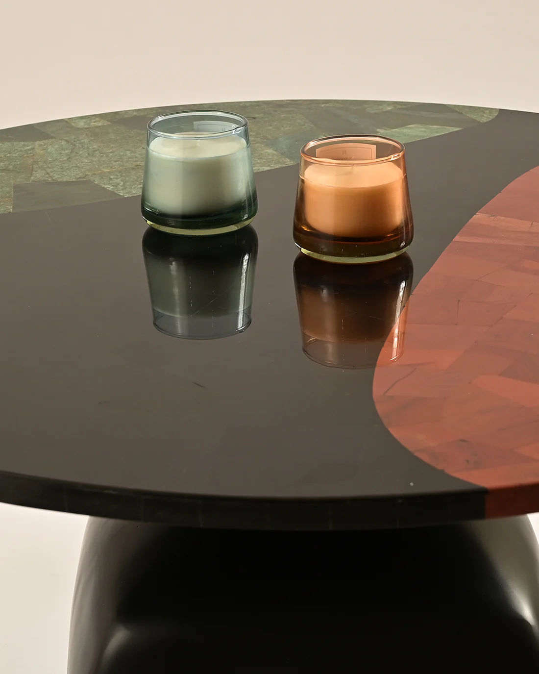 LMNOH's Jasper Coffee Table features a modern design with geometric green and reddish-brown patterns. Its cast aluminum base adds elegance, complemented by two small candles on the tabletop: one white in a blue holder and another orange in clear.