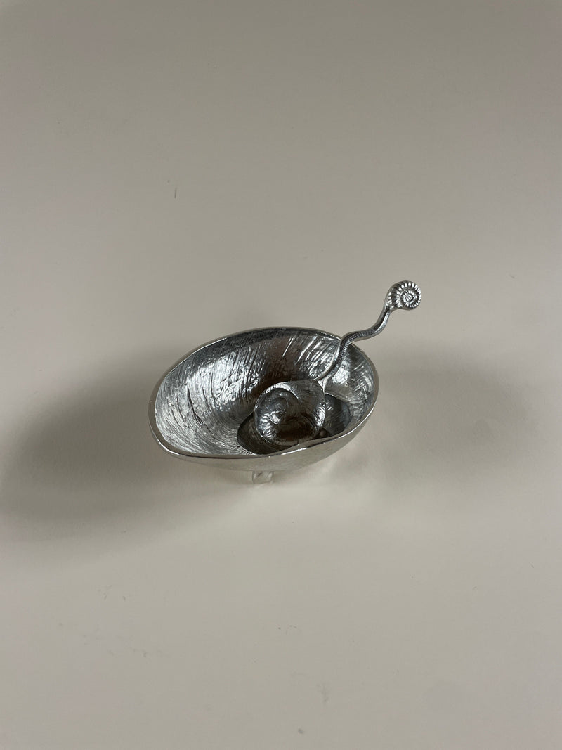 A metallic, bowl-shaped sculpture crafted by Maud Vaughan, known as the Shell Dish & Spoon, rests on a neutral surface. Its twisted, ornate handle bears an intricate seashell-like design that harmonizes with the vintage polished pewter bowl's spiral element at the end. The textured surface is accentuated by strategic lighting.