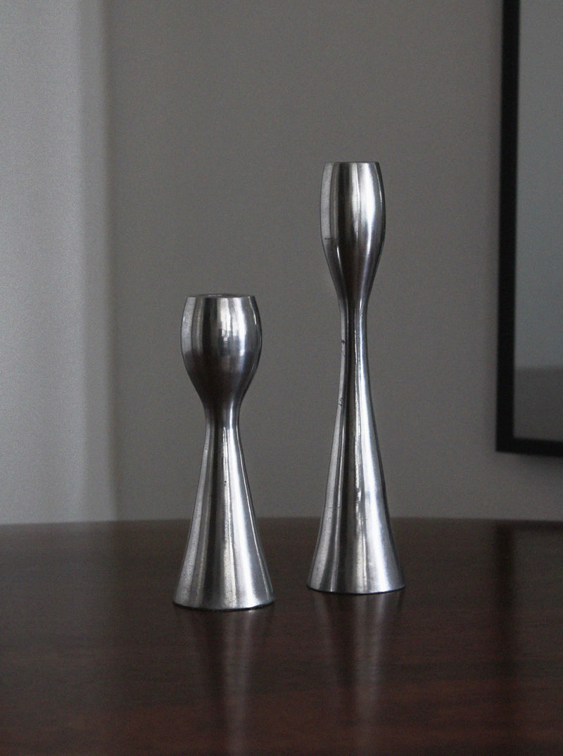 Two Polished Aluminum Candleholders from Vintage Stories, each a different height, rest on a dark wood surface with part of a framed mirror in view—a stylish accent for any living room.