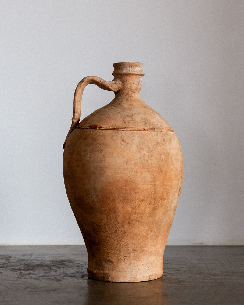 An Early 20th Century European Olive Jar by Spigel, featuring a narrow neck and single curved handle, stands on a plain surface. Its rustic textures and warm brown hues evoke the charm of hand-crafted pottery against a neutral background.
