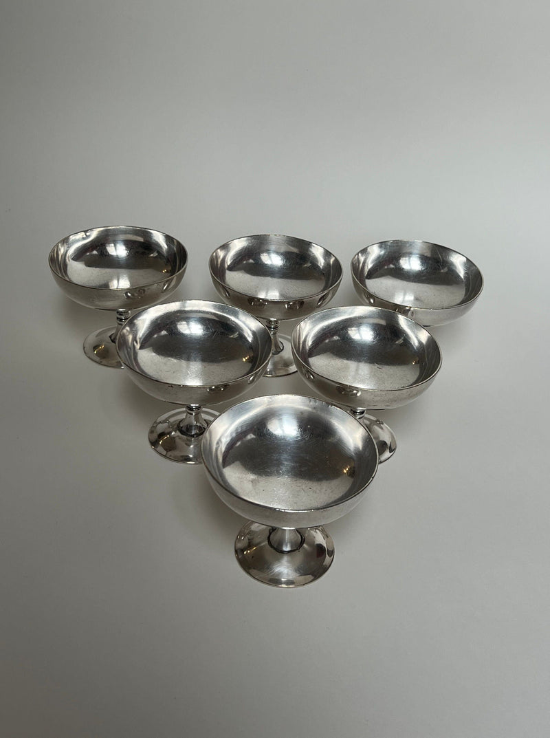 Six Heavy Silver Coupes by Manushi, featuring wide, shallow tops and pedestal bases, are arranged in a triangular formation on a plain light-gray background.
