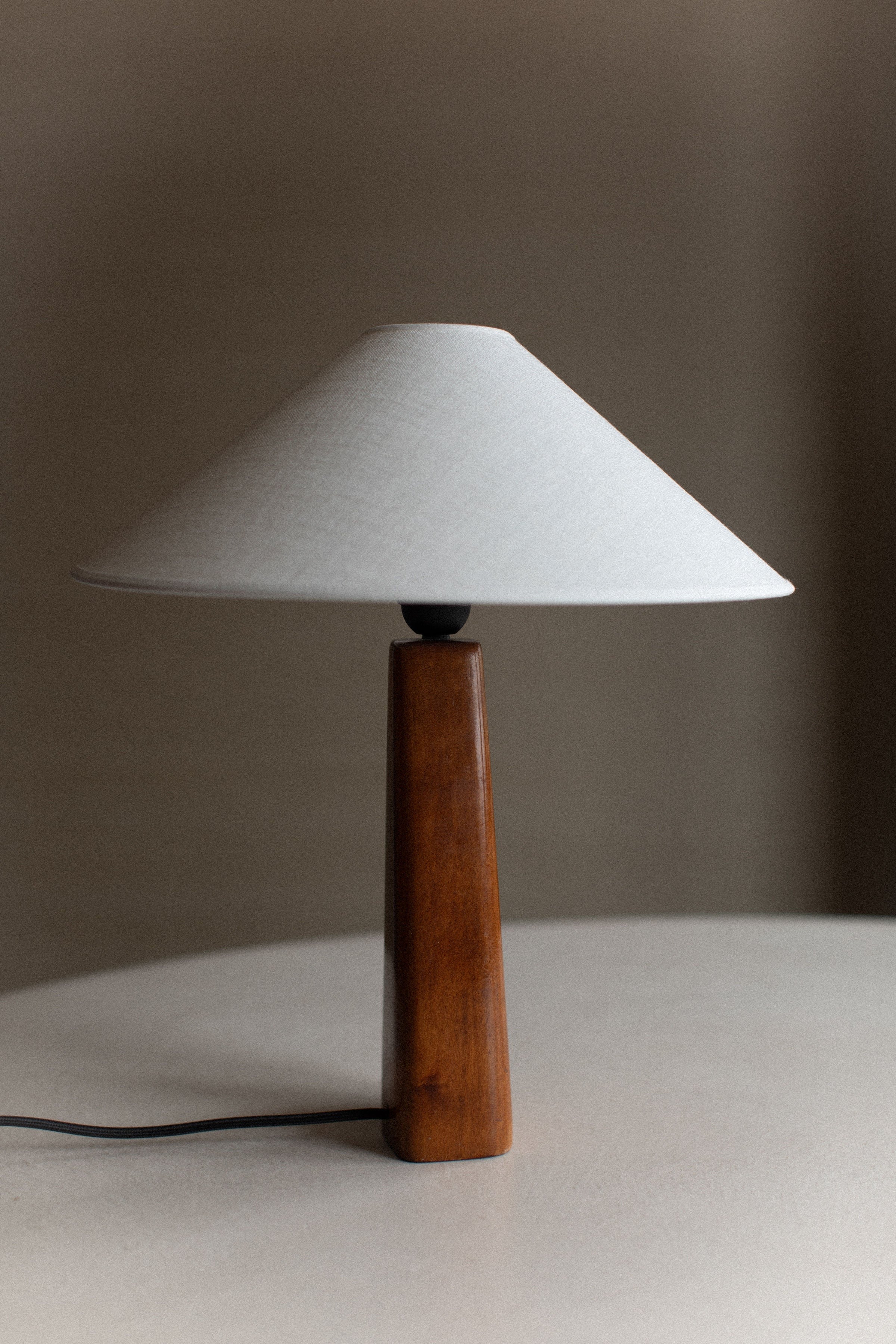 A minimalist table lamp with a conical white shade and a wooden triangular base with hand-carved details sits on a light grey surface. The simple, modern design features an elegant contrast between the light shade and dark wood. The 1960's Wooden Lamp by Out For Lunch includes a black power cord that extends from the base.