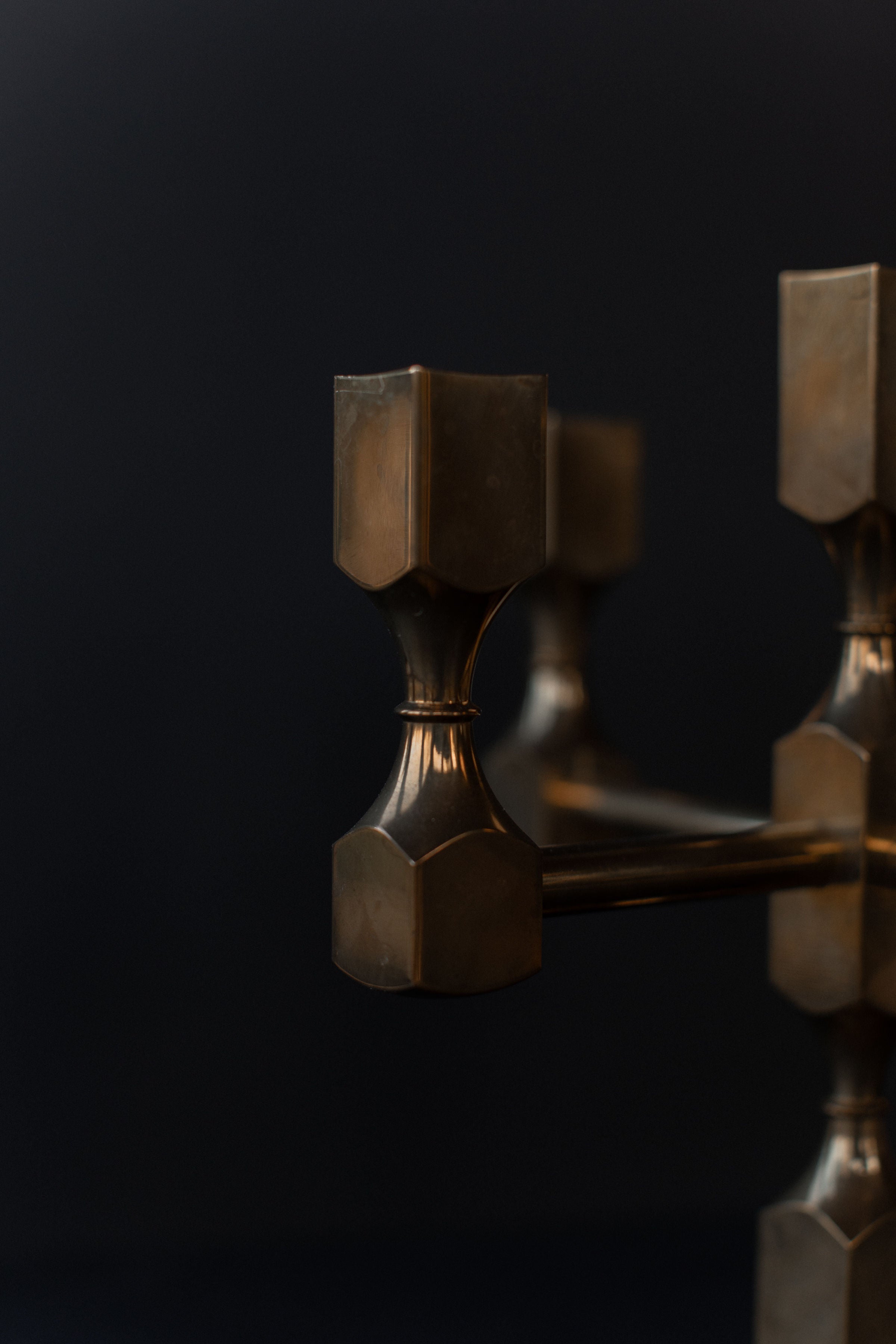 The Brutalist Brass Candleholder by Out For Lunch showcases cylindrical and hexagonal shapes on a reflective bronzed surface, set against a dark background. Its solid brass construction enhances its industrial aesthetic, making it both functional and decorative.