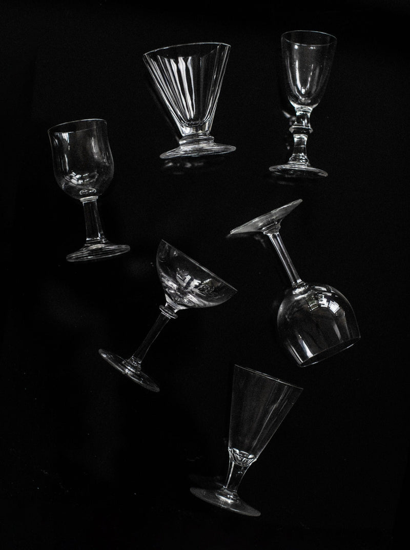 A set of six mix-matched glasses by Out For Lunch, featuring diverse types of empty glassware such as wine glasses, cocktail glasses, and goblets, are arranged diagonally against a dark background. Each curated piece is oriented in various directions, creating an artistic and dynamic composition for a sophisticated drinking experience.