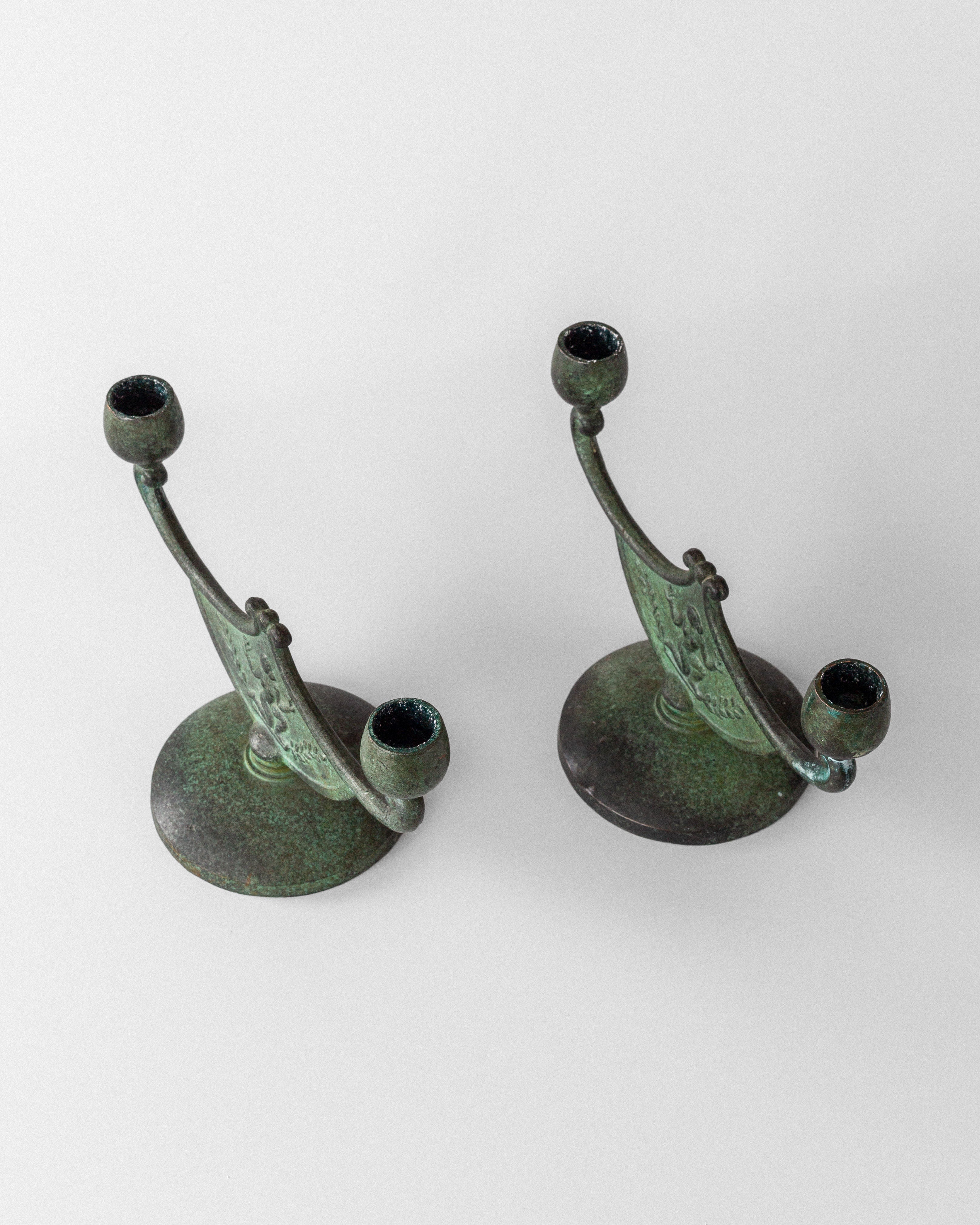 The Spigel Pair of Art Deco Bronze Candleholders features an antique, oxidized finish with ornate designs and a green patina. They stand against a plain, light background.