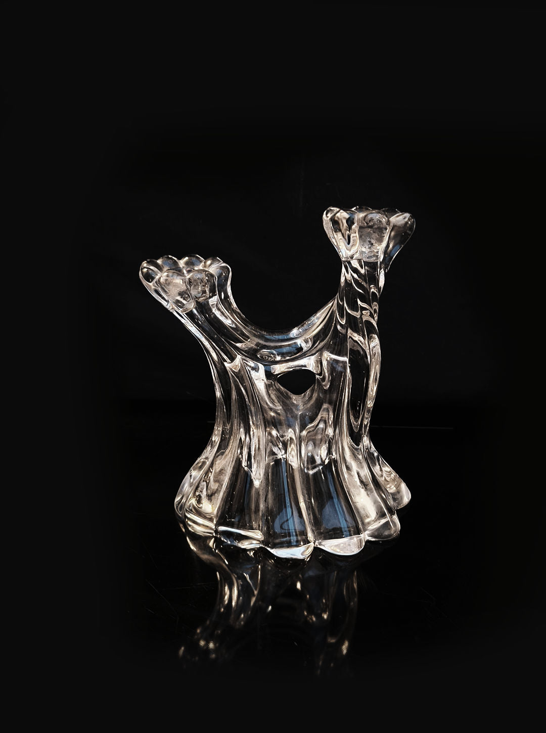 A transparent, sculptural glass candle holder from Les Objoies stands against a dark background. Its wavy, flowing design and textured surface evoke twisting waves or molten glass, giving it an artistic and elegant appearance.