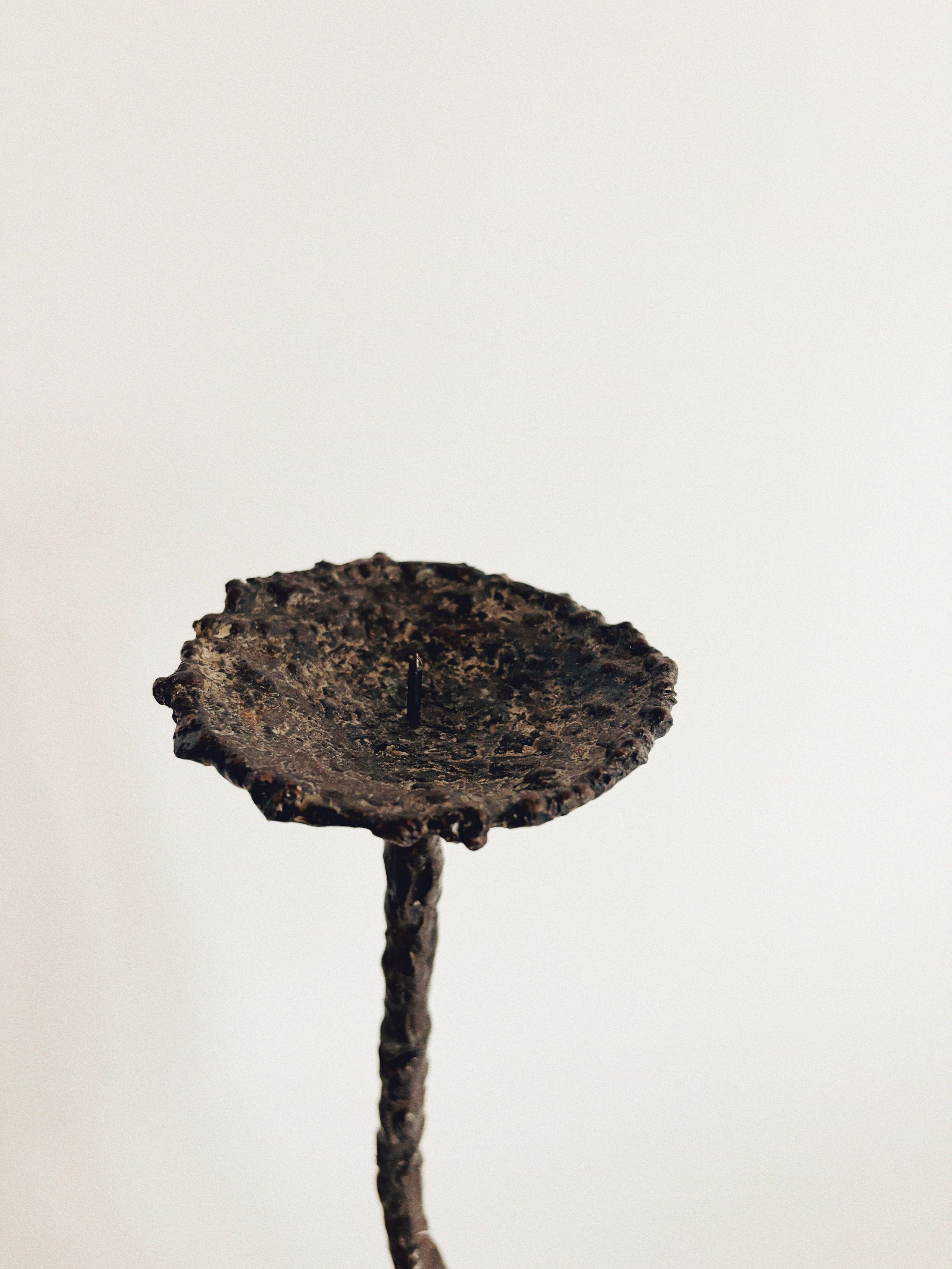 A detailed close-up of the Molten Metal Artisan Candlestick by Huerto highlights its aged, textured finish and molten metal origins, featuring a round, dish-like platform atop a slender, slightly twisted stem. Its artistic form is prominently displayed against a simple, light-colored background.