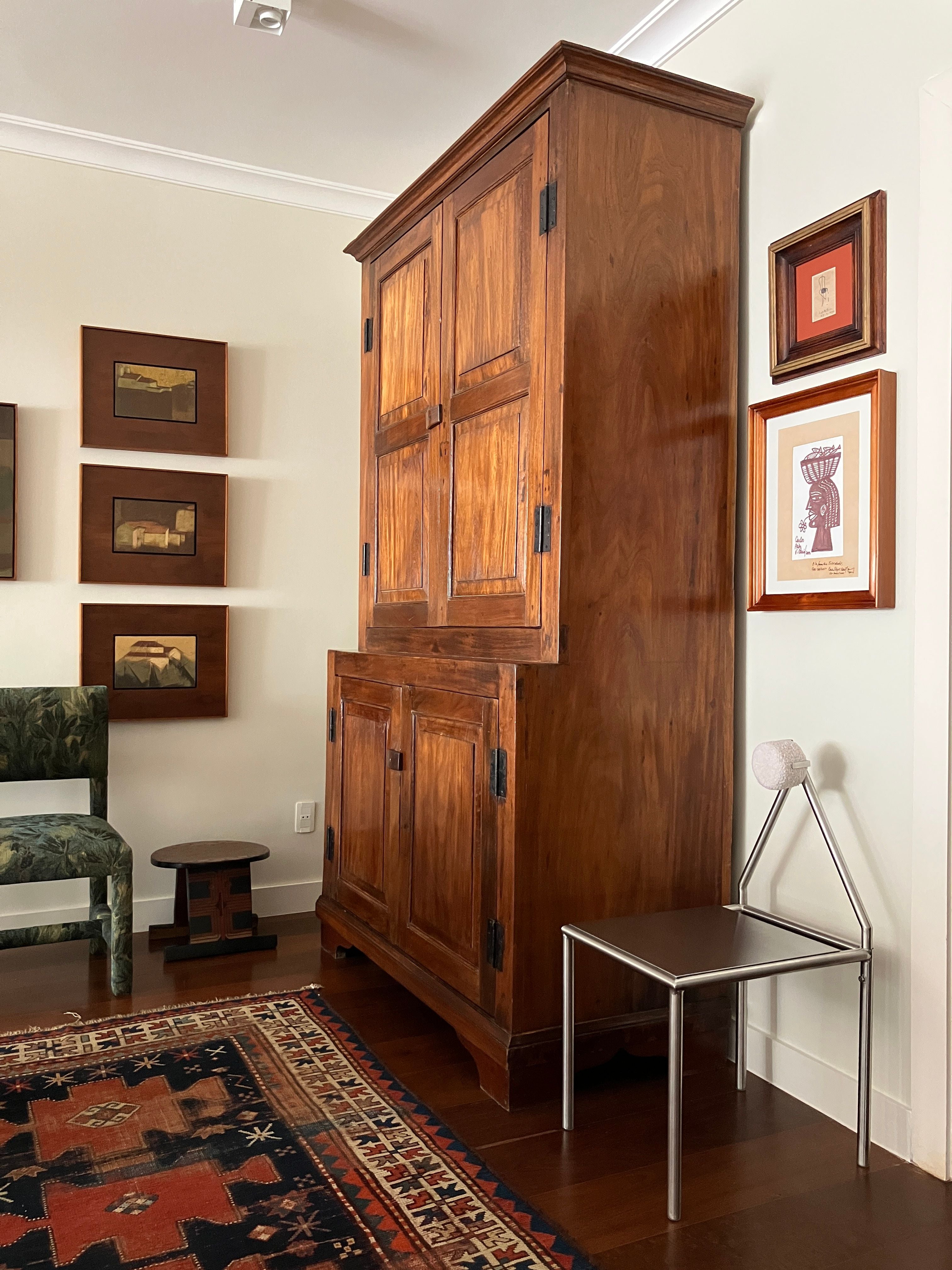A wooden cabinet stands against a light wall, surrounded by framed art, a green chair with removable cushion, a small round stool, an HT. 'H01' Stainless Steel Chair, and a patterned rug on the wooden floor.