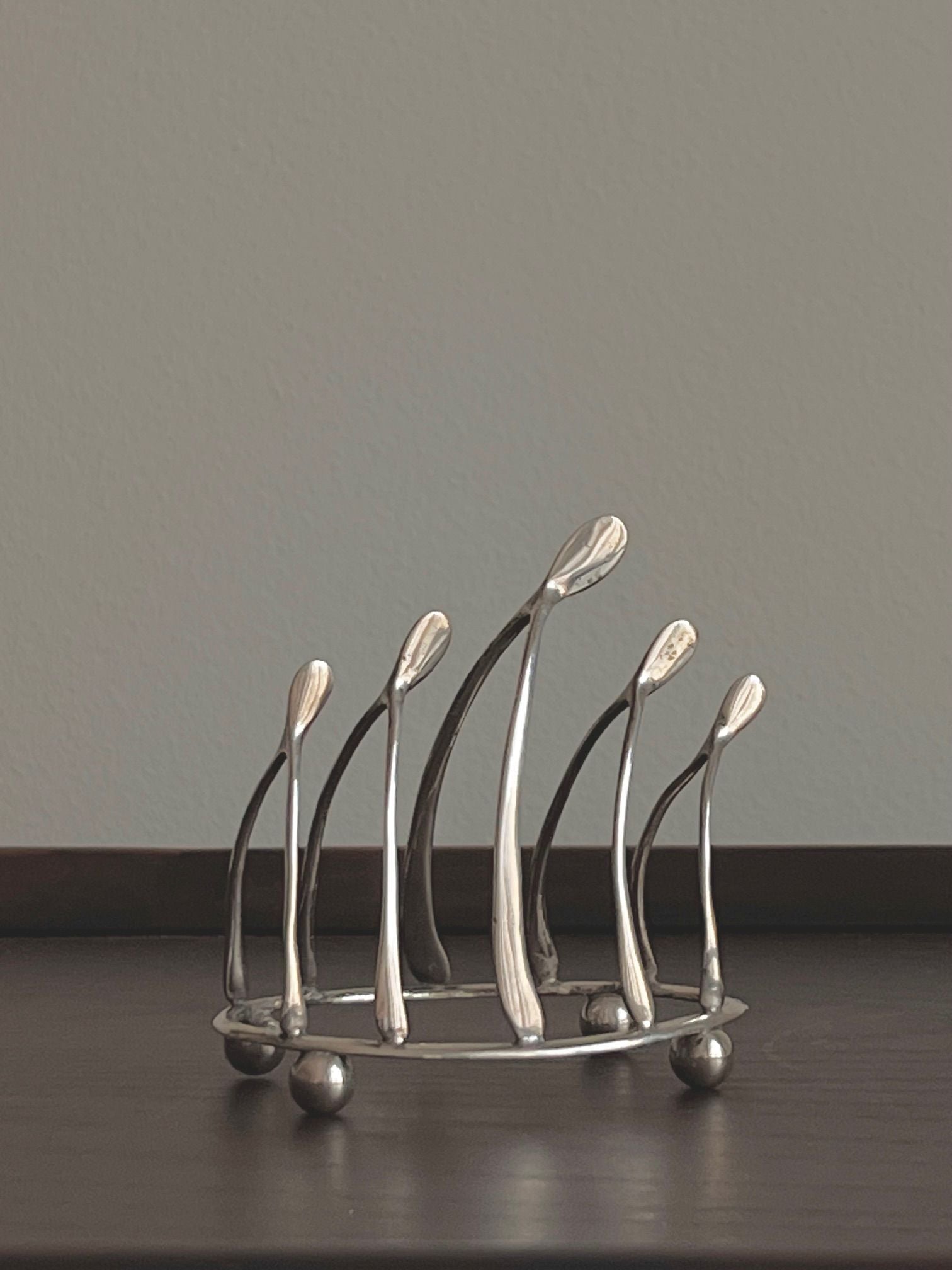 A metal Wishbone Silver Toastrack by Médecine, with elongated abstract spires, rests on a dark wooden surface. Its reflective silver finish and minimalist design contrast elegantly against the plain, light-colored wall, resembling the work of an English silversmith.