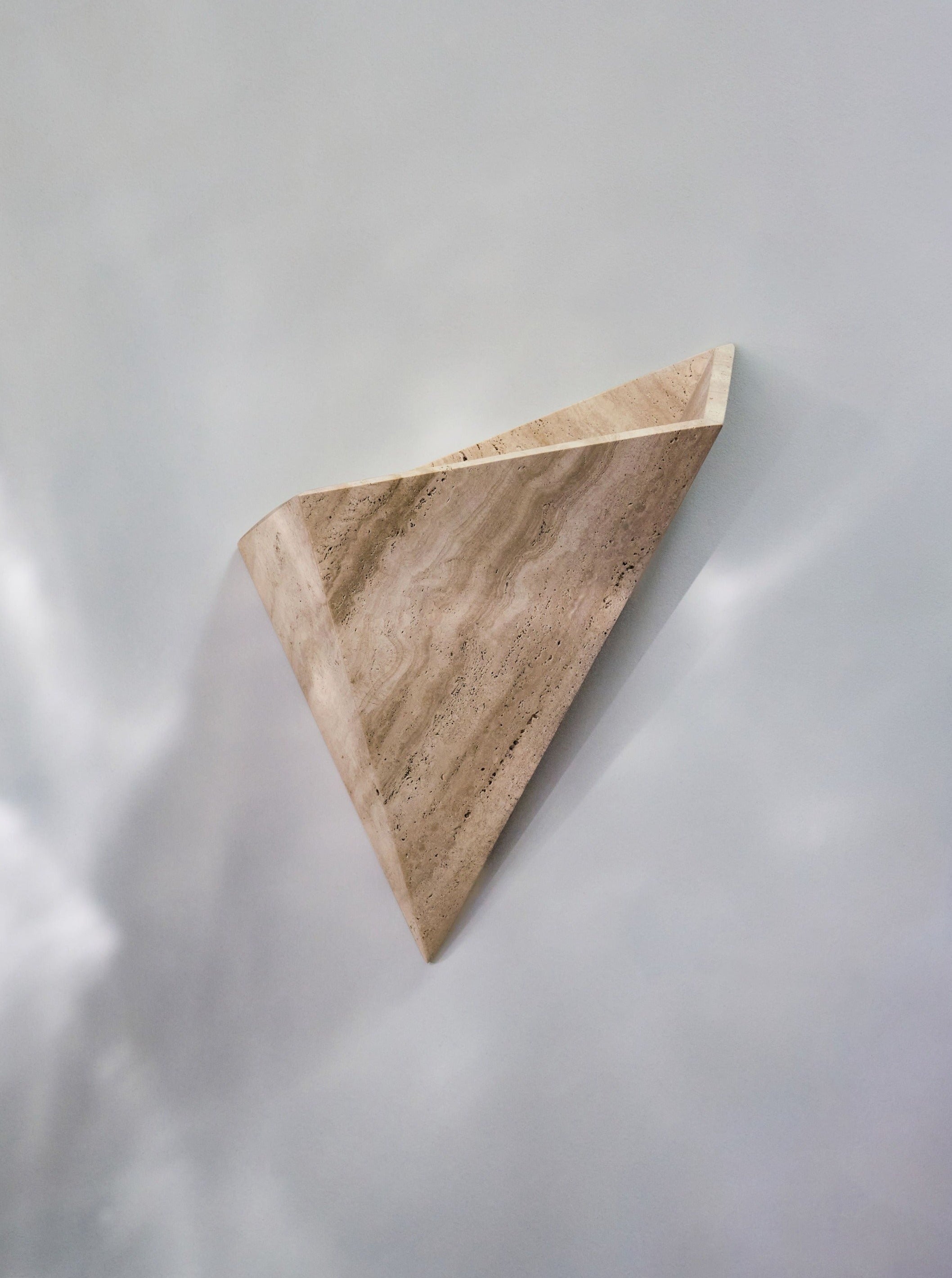 A triangular piece of polished travertine mounted on a light gray background, creating soft shadows around its edges.