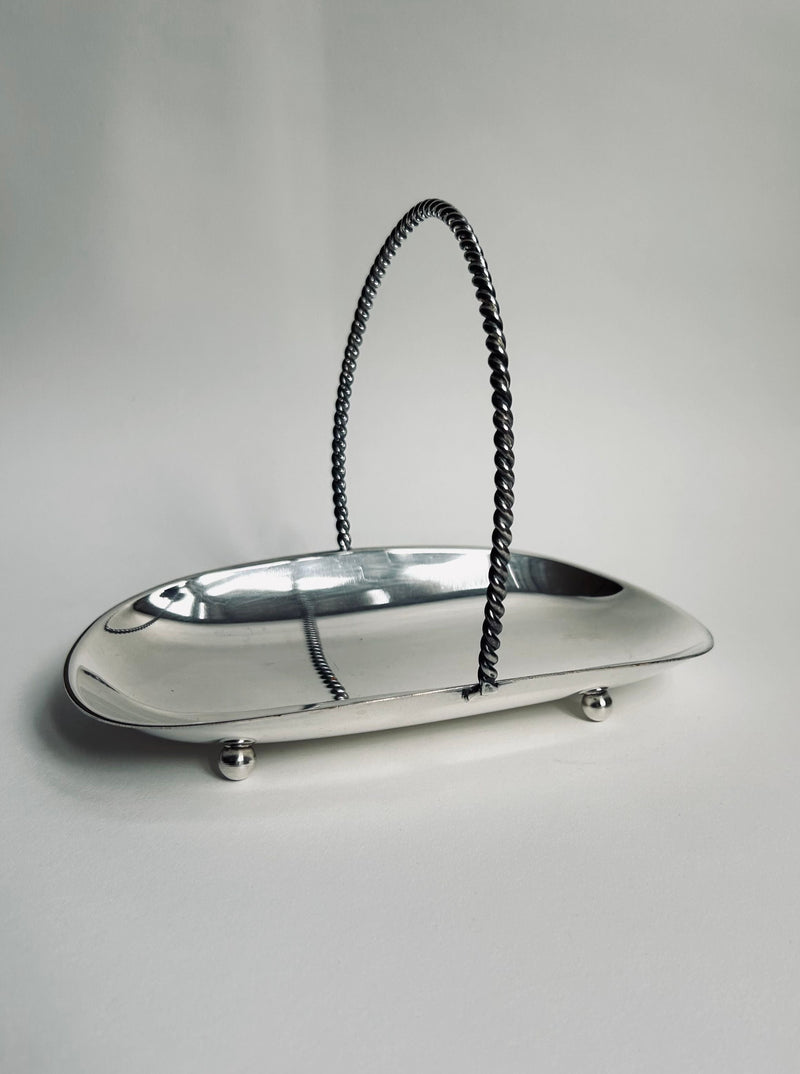 A polished Art Deco Silver Plated Basket by Flock Studio features a rectangular design with a twisted handle and four rounded feet, set against a plain background. The basket's reflective surface exudes elegant Art Deco charm, suggesting both decorative and functional uses.