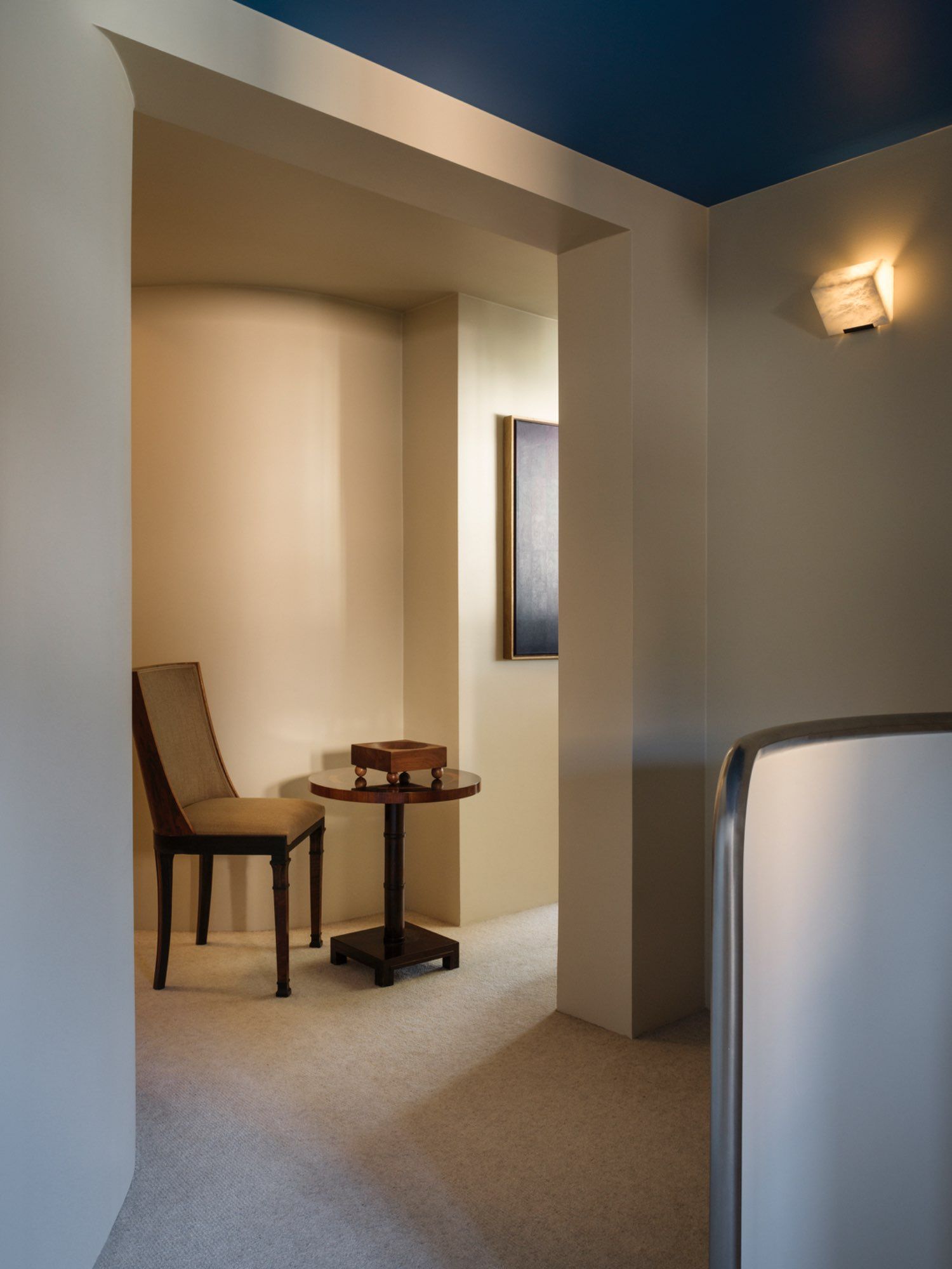 A minimalist room with beige walls features a wooden chair and a small round table, adorned with the Tray 01 Walnut from Studio Vraco. A framed artwork graces the wall, while a modern wall sconce casts warm light. The ceiling is painted in dark blue, enhancing the contemporary design.