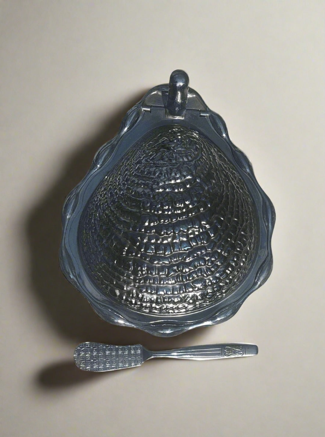 The Les Objoies Shell Butter Dish, shaped like a vintage silver clamshell and adorned with intricate textured details, is placed on a marble surface. Below the dish rests a small silver spreader featuring a handle with a matching textured design. The shadows of the items are cast on the background.