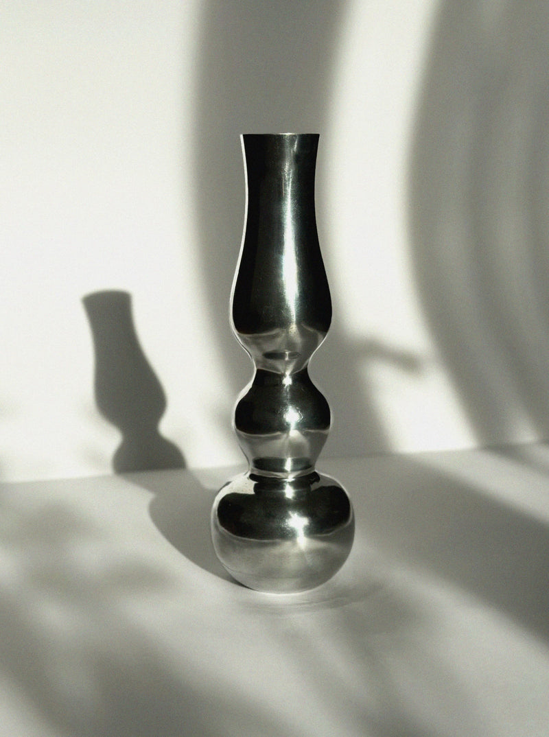 The Aluminum Figure Vase by Vintage Stories features a tall, metallic form with a mirror finish and sculptural curves. Set on a white surface, it casts bold shadows while blurred branches in the background highlight its minimalist elegance.