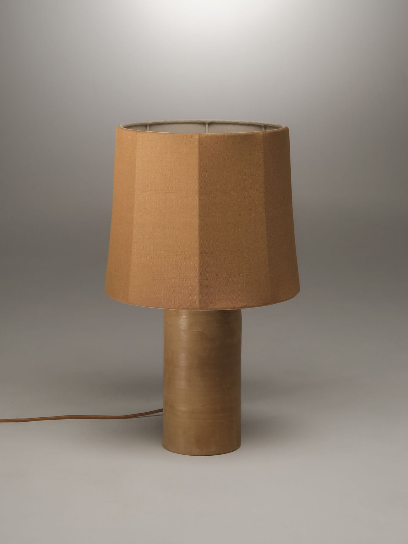 A simple, modern table lamp with a terracotta base and a drum-shaped, beige lampshade made from pure linen. The Sonora Linen Table Lamp by Nassi Lamps is turned off and placed against a plain, light-colored background. A power cord extends from the base of this stylish Puglia-inspired piece.