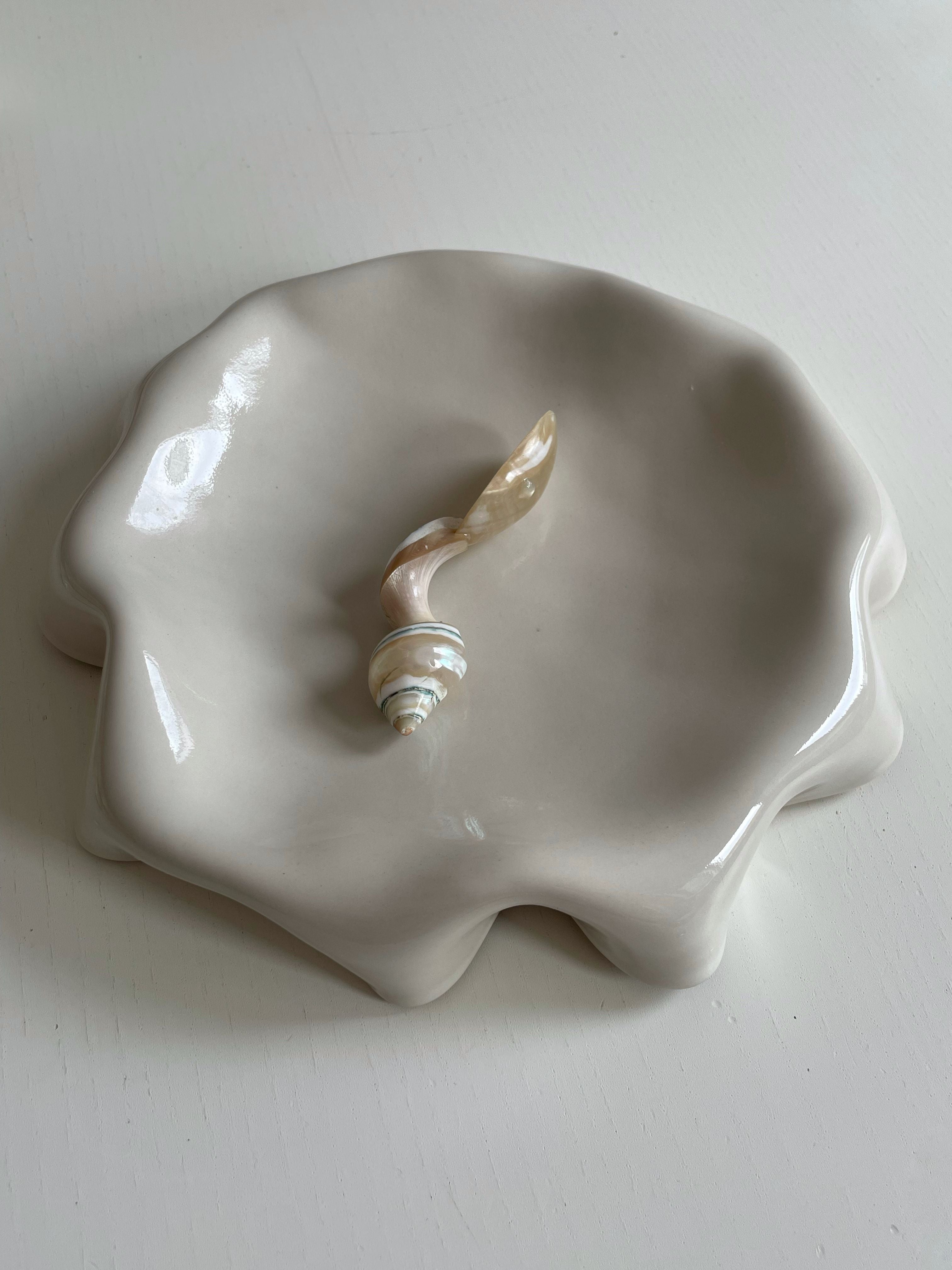 The Melted Butter Plate in Off White by LESS, a handmade stoneware ceramic dish coated in a glossy off-white glaze and designed to resemble a melted form on a white surface, holds a small, decorative object that looks like a mix of a seashell and a leaf.