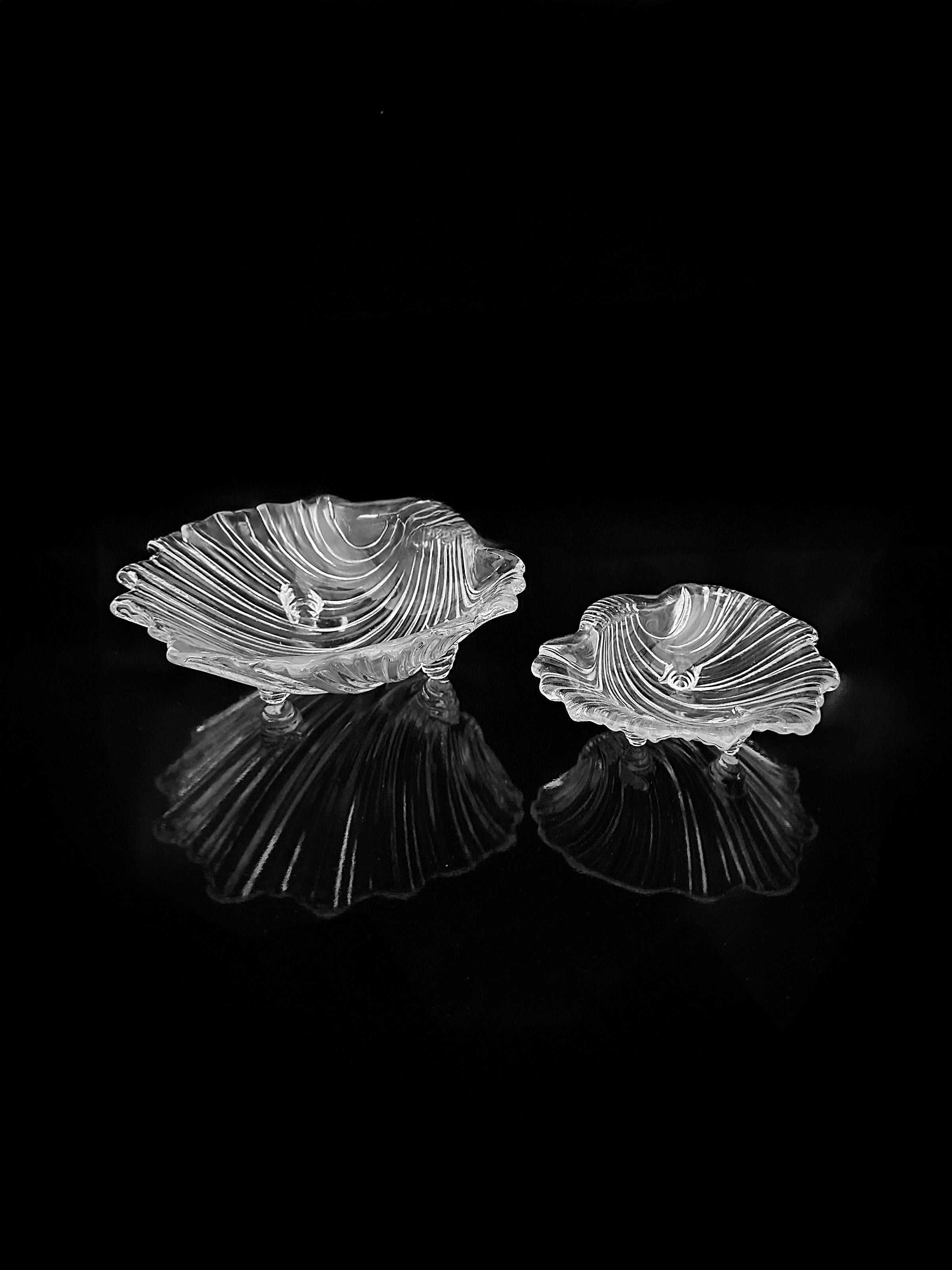 Two "1980s Shell Shaped Glass Bowls" from Dodo Vintage, featuring an exquisite design, display intricate patterns against a black background. The larger bowl is positioned on the left and the smaller one on the right, perfect for presenting delicacies. Both bowls elegantly reflect subtle light.