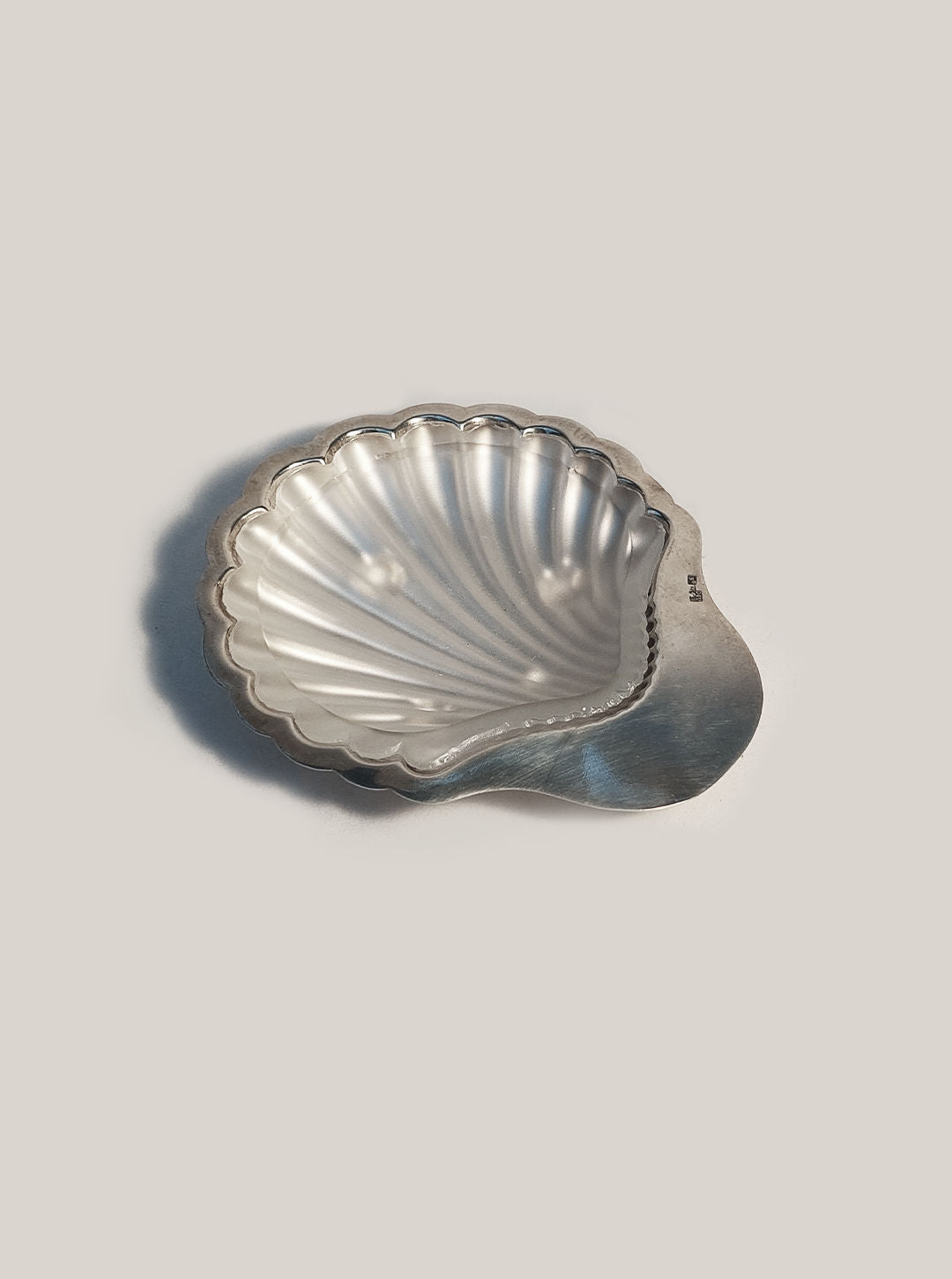 The Shell Butter Dish by Les Objoies, a shiny, silver-plated scallop shell with a ridged texture, sits on a plain, light background.