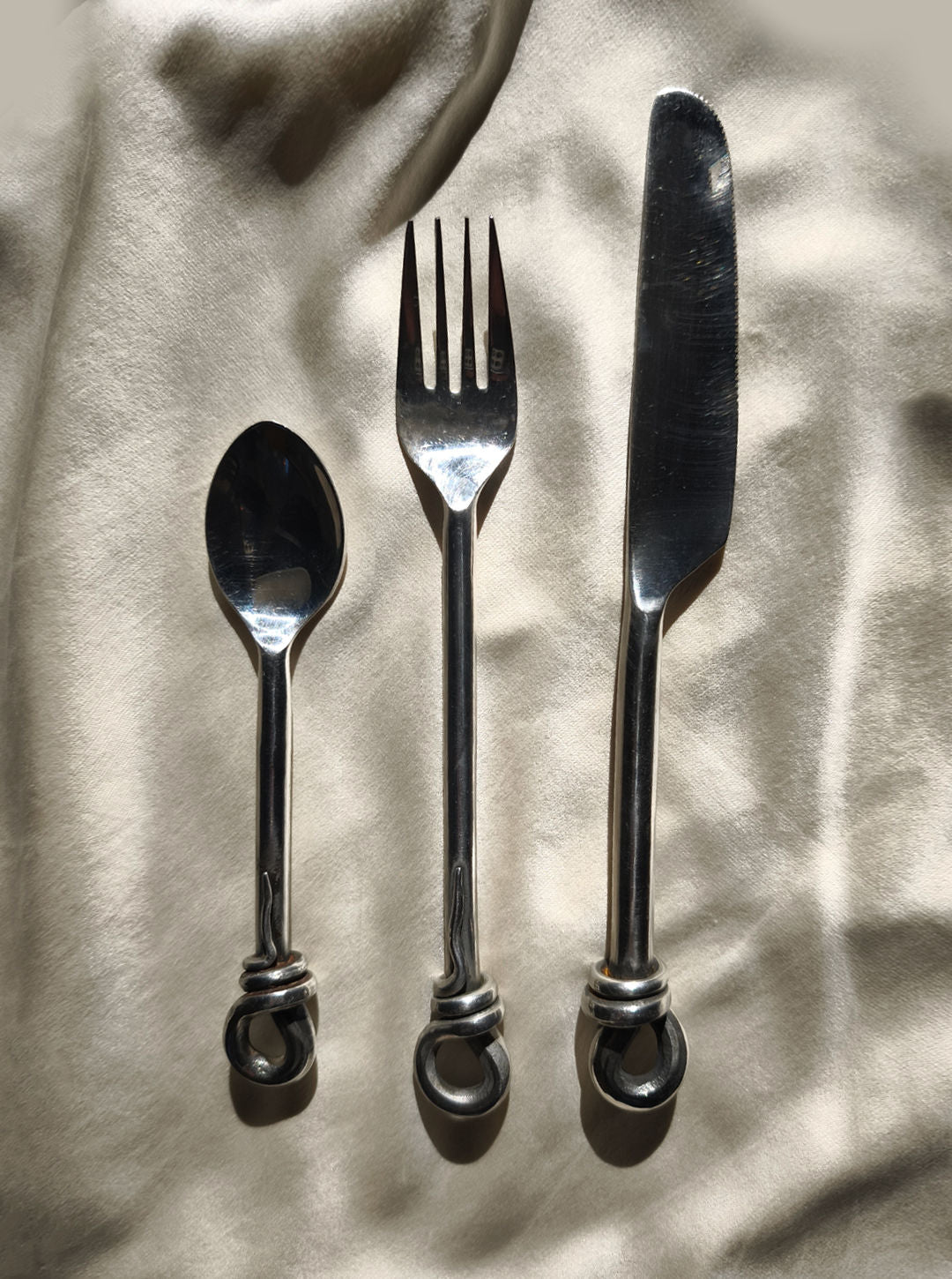 The Les Objoies Set of 3 Twist Cutlery, featuring a spoon, fork, and knife with uniquely twisted handles and an organic shape, is elegantly arranged side by side on a soft, light-colored fabric under gentle sunlight.