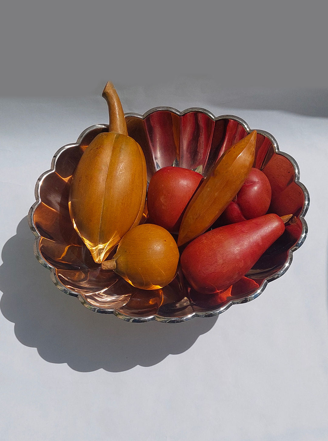 The Large Fruit Basket by Les Objoies, showcasing an elegant scalloped metal design, is filled with a variety of wooden fruits, including a pear, an apple, and a gourd. Set against a plain background with soft lighting and subtle shadows, it makes for the perfect decorative centerpiece.