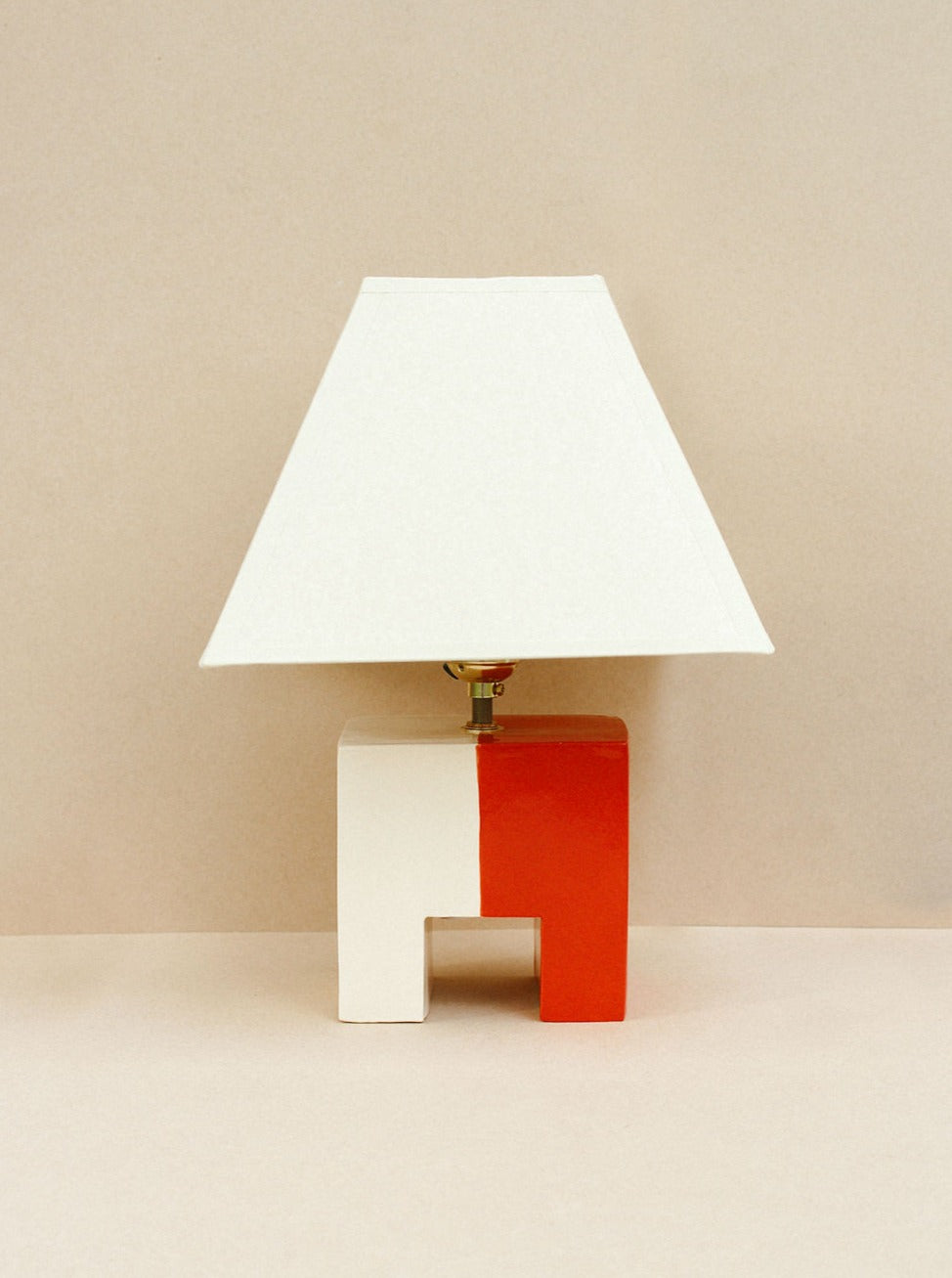 A modern hand-built ceramic Cristo Lamp (Rojo/Hueso) by Casa Veronica with a geometric base divided diagonally into red and white halves. It features a simple, custom square lampshade and is set against a beige background, ensuring both style and safety as it's UL listed.