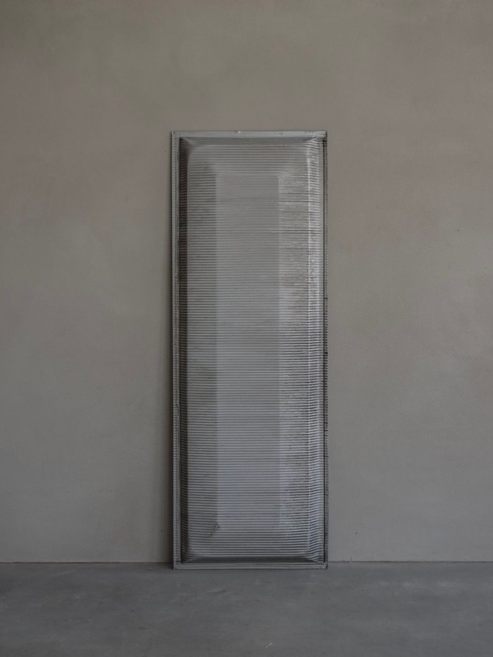 A tall, rectangular Jean Prouvé - Façade Panel 70 by Bicci de' Medici stands against a gray wall on a concrete floor. The ribbed texture and slight distortion effect create an intriguing visual ripple reminiscent of modern designs.