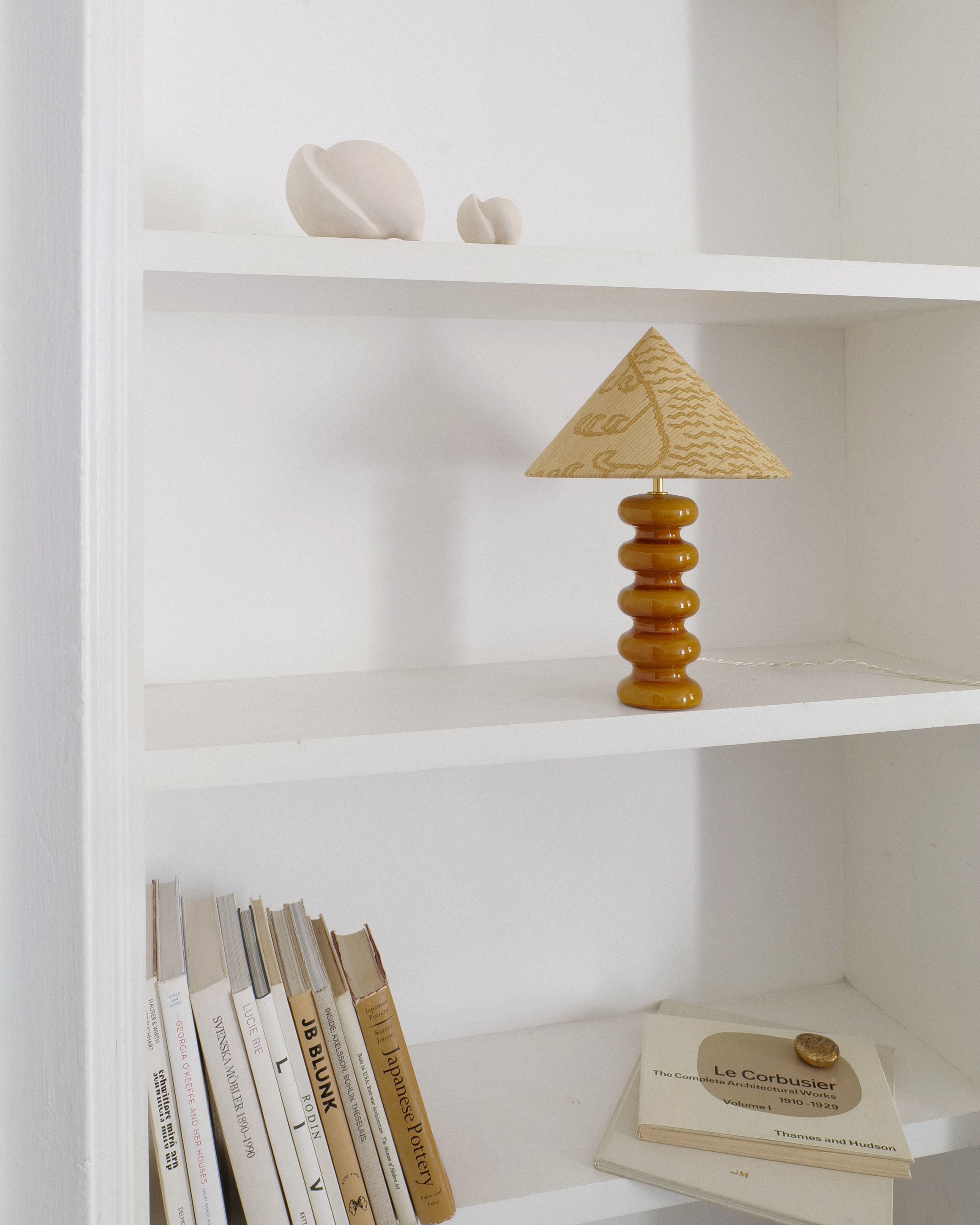 A minimalist interior features white shelves with a wavy wooden lamp by Collection apart and a Sculptural Ceramic Lamp with a beige shade. Books are stacked below, while the top displays smooth abstract ceramic pieces reminiscent of Doulton England, alongside an open "Le Corbusier" book.