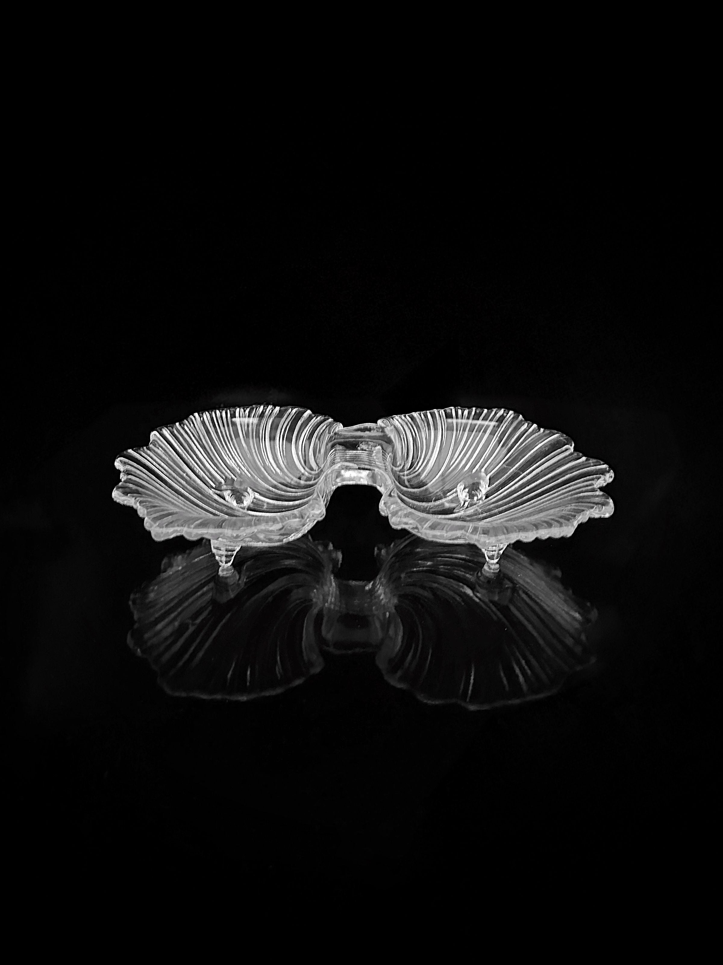 A set of beautifully crafted, 1980s Shell Shaped Glass Bowls by Dodo Vintage features intricately designed clear glass that resembles seashells, elegantly connected at the center and set against a black background. The symmetrical reflections highlight their graceful curves and intricate details, making them perfect for stylishly showcasing delicacies.