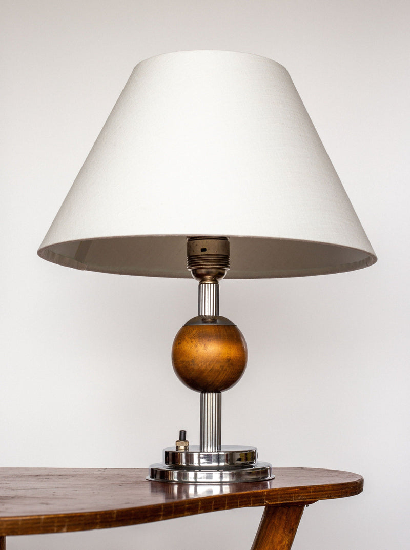 The Spigel Swedish Art Deco Metal and Wood Lamp, 1930s features a white conical lampshade, paired beautifully with chrome and wood elements. This handcrafted design from the 1930s includes a wooden spherical accent in the center and a metal base with a switch. This elegant lamp is set on a wooden surface against a plain background.