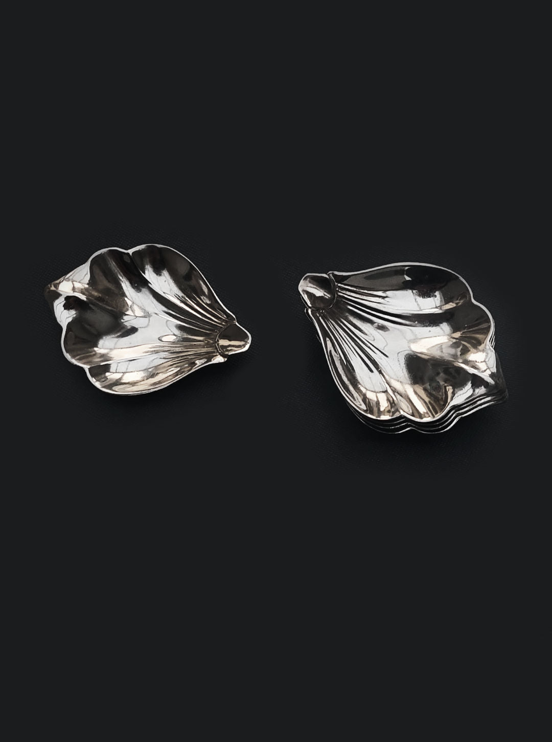 Two elegant pieces from the Les Objoies Set of Petal Dishes are artfully displayed against a dark backdrop. These silver, leaf-shaped dishes boast intricate detailing and a reflective surface that beautifully captures the light, showcasing their sophisticated design.