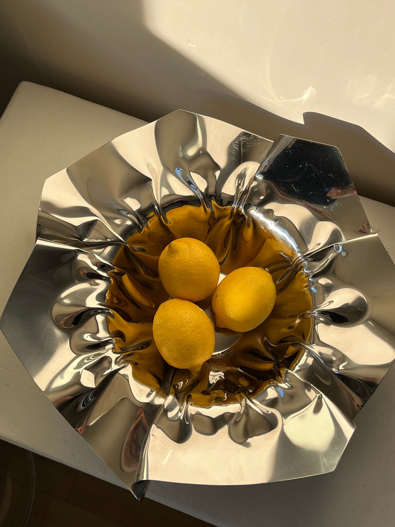Three lemons rest in a shiny, crumpled metallic bowl from septembre studios called the Folded Chrome Bowl, reflecting natural light. The bowl’s unique texture and design create dynamic patterns of light and shadow around the yellow fruits, adding an artistic touch to the interior decor.