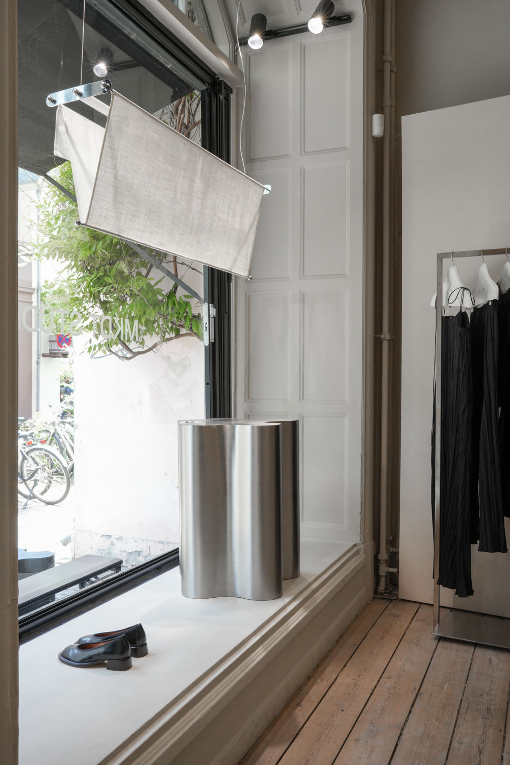 A minimalistic store display features the Vento Pendant by Caia Leifsdotter, a large stainless steel vase, black sandals, and black pleated clothing on an adjustable rack, set by a window with modern neutral decor and light wooden flooring.