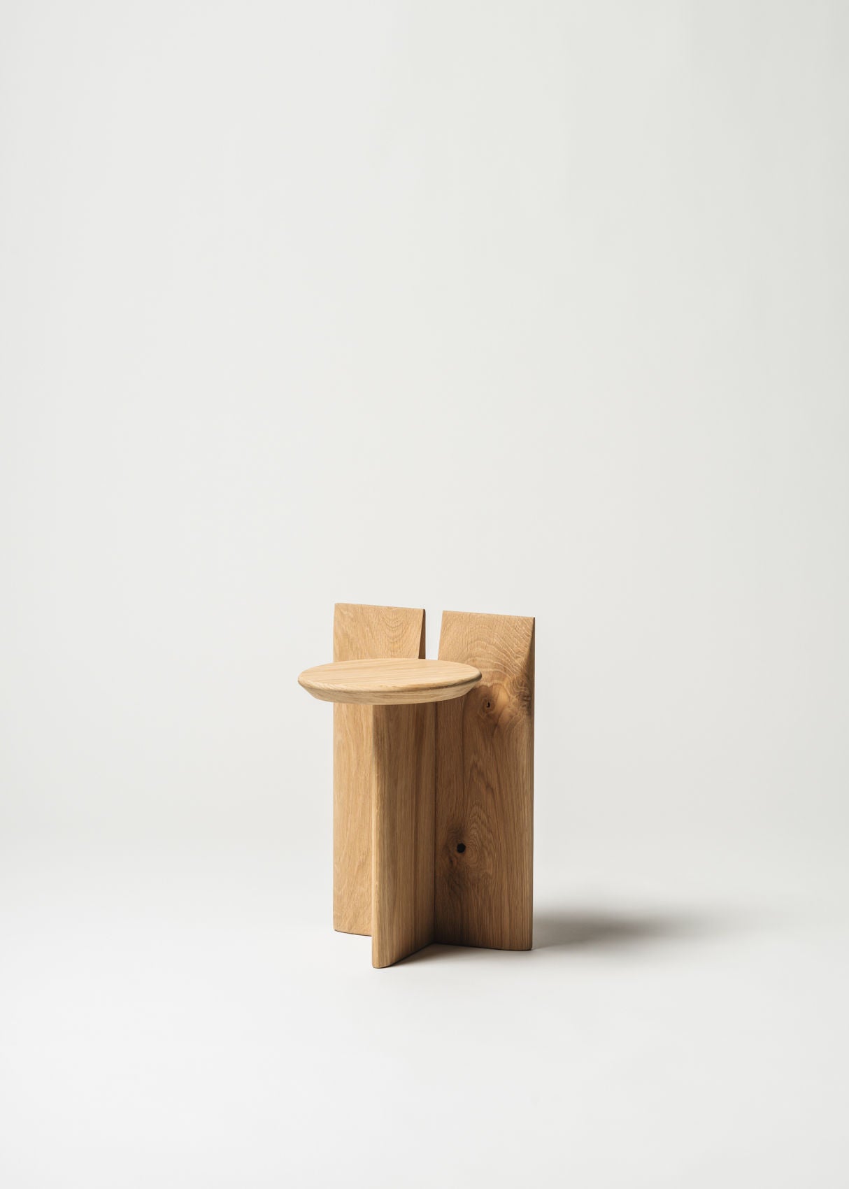 The Tabi Stand chair by Obstacles is a minimalist piece made from light oak. It features a geometric design with a flat round seat and two rectangular back panels, all against a plain white background, emphasizing its modern aesthetic and natural materials.