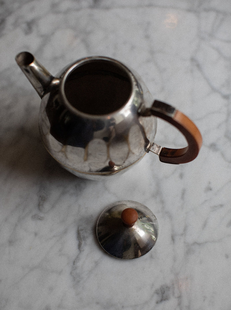 A silver-plated Small Teapot by Out For Lunch with a wooden handle and matching lid is placed on a white marble surface. The teapot has an open spout and a reflective finish. The lid is situated beside the teapot, exposing its interior. This timeless elegance serves as a stunning decorative accent.