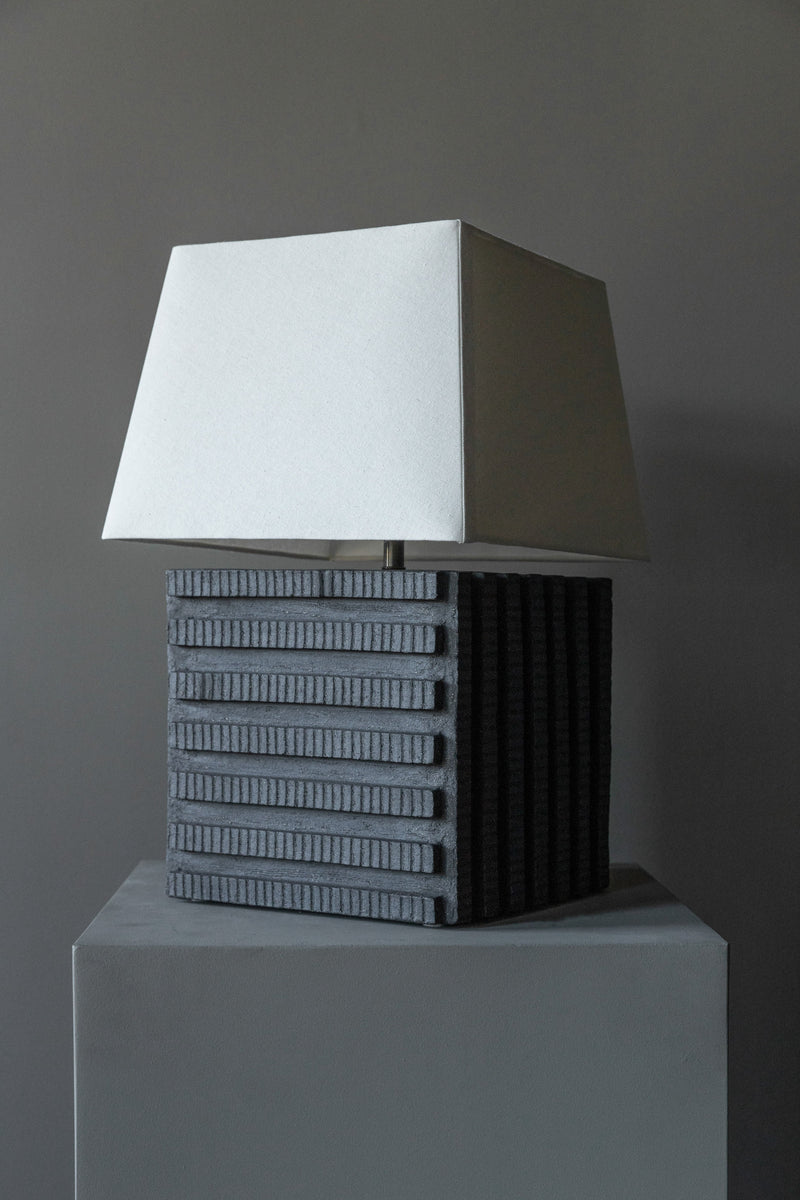 The Akune Ceramic Lamp - Number One by Studio OSKLO, with a rectangular white shade and dark geometric base inspired by post-Bauhaus brutalism, is displayed on a gray pedestal against a plain gray background.