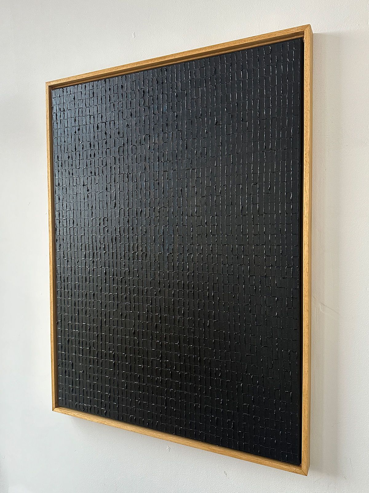 Displayed on a white wall is "Lamp Black," an oil on canvas artwork by Roi Elmaliah, featuring a textured black surface adorned with a gridded pattern. This piece, influenced by the vibrant arts culture of Amsterdam, boasts a wood frame that provides striking contrast to its dark tones.