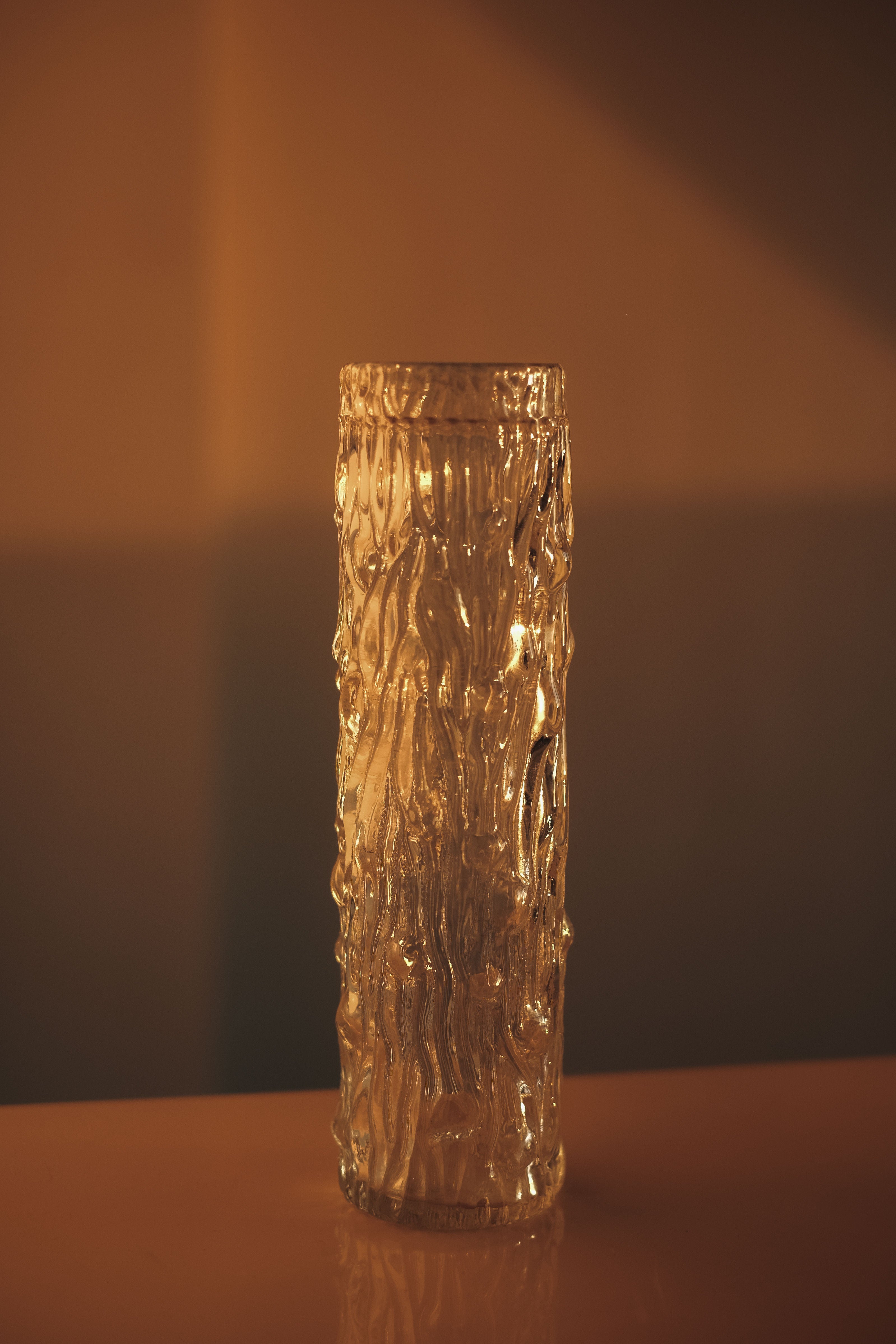 A Small Glass Cylinder Vase by septembre studios, reminiscent of a Sklo Union design, holds a lit candle that casts a warm, soft glow. The gently blurred background emphasizes the cozy ambiance created by the candlelight.