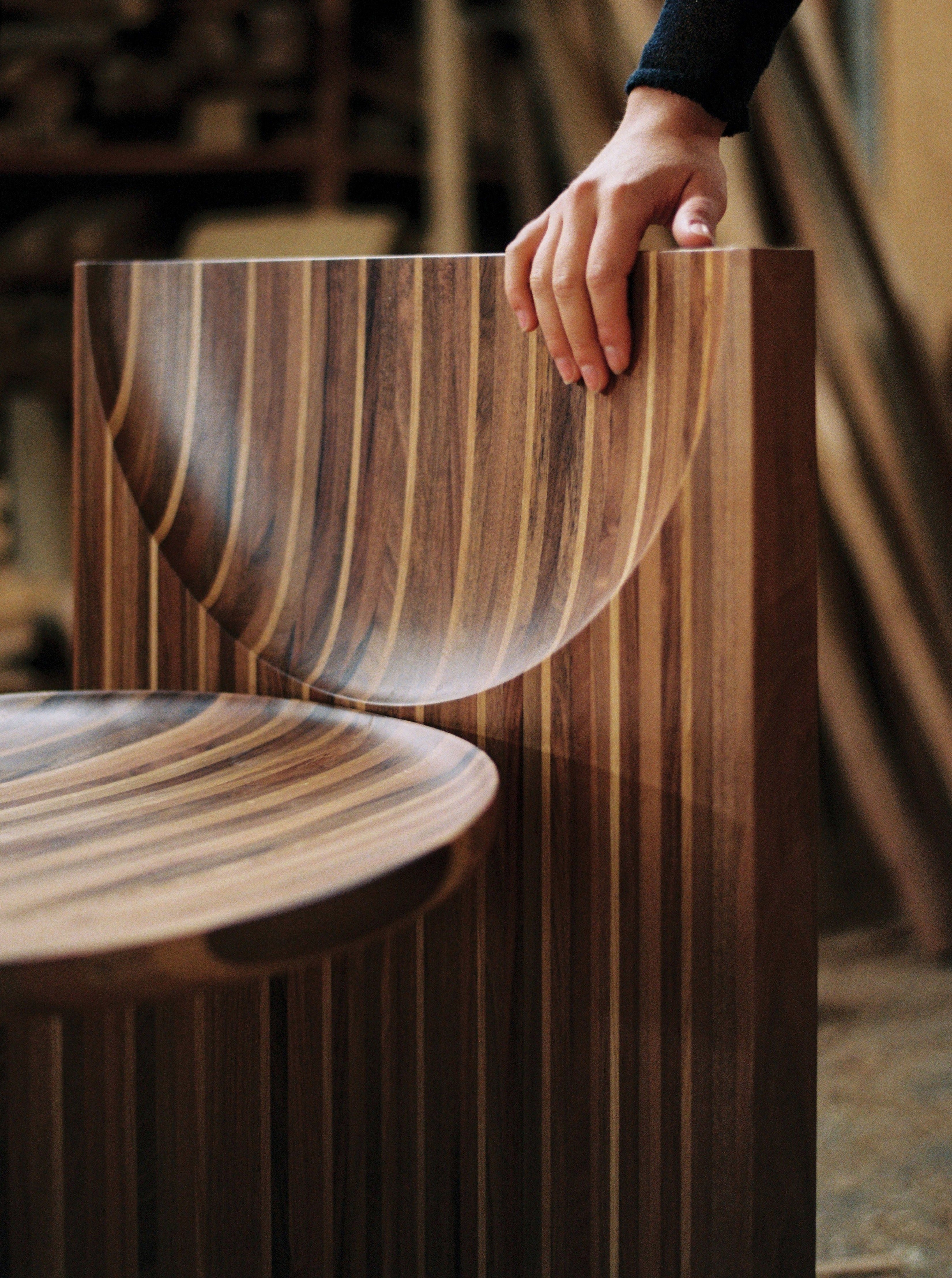 A hand rests on the back of the Vaga Chair by Rosana Sousa, a modern wooden piece featuring an intricate striped pattern. The solid walnut seat and backrest showcase smooth, curved surfaces, set against a backdrop of blurred elements from a workshop environment.