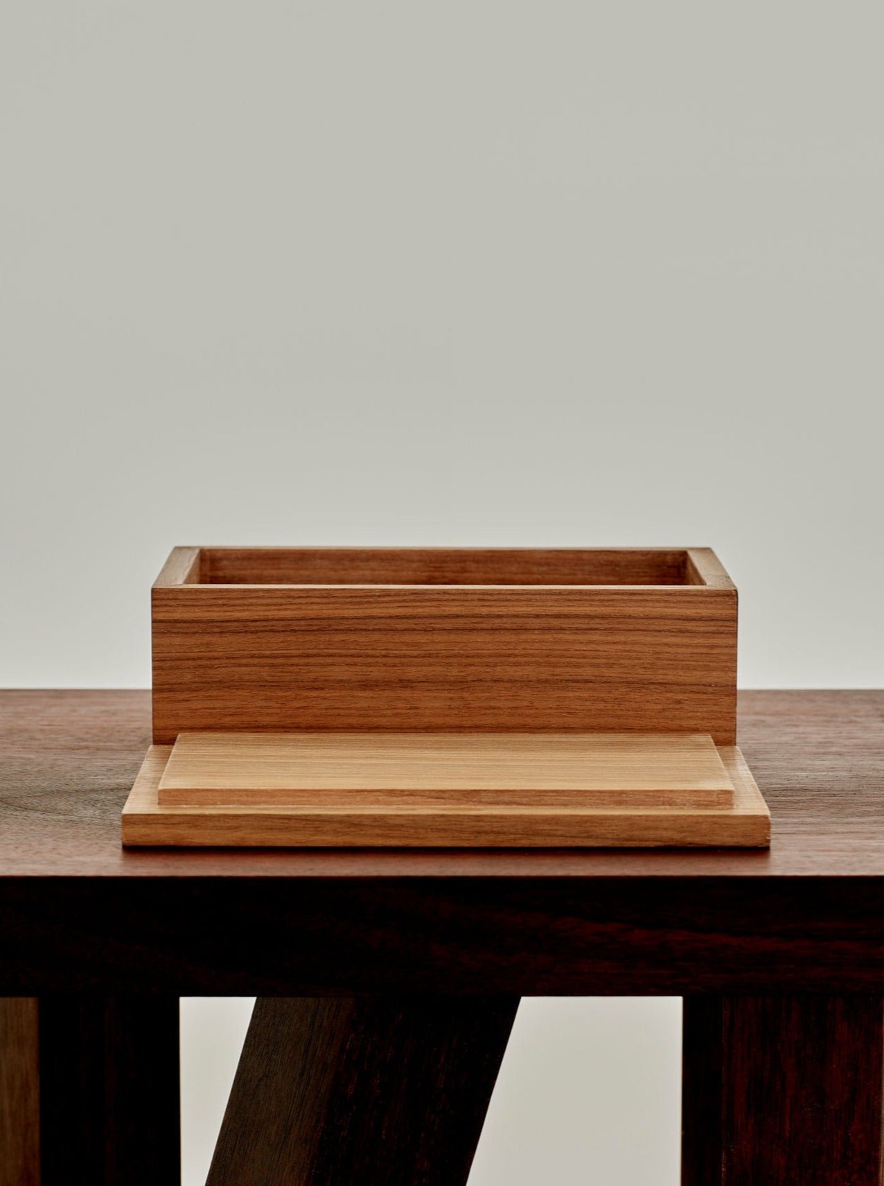 A MOCK STUDIO Miter Box with an open top sits on a square wooden table. The table has a dark finish with sturdy, angular legs. The MOCK STUDIO Miter Box is smaller and has a lighter wood finish, placed centrally on the table against a plain, light background.