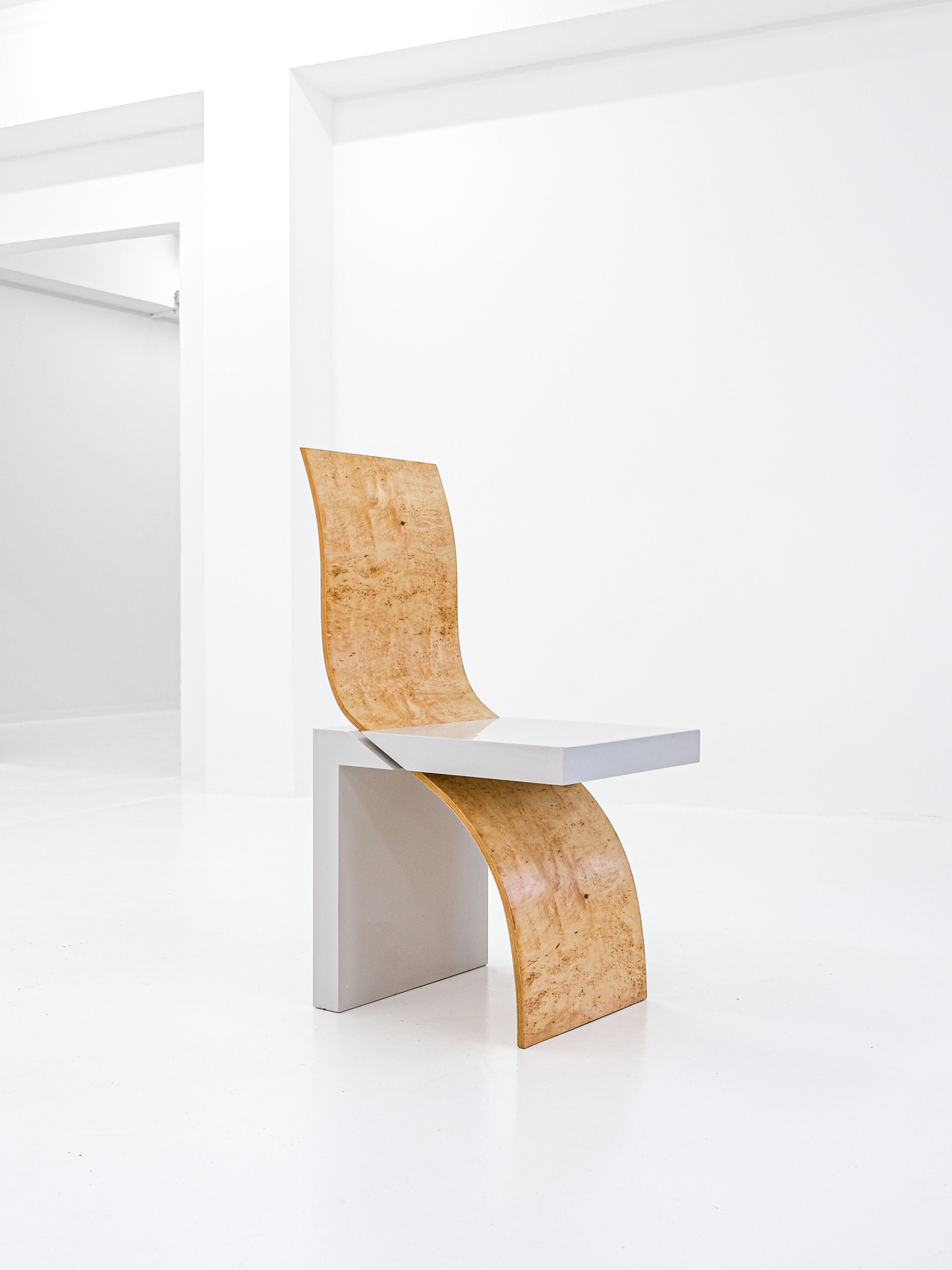 The Postmodern Leda Chair by Angela Oedekoven for antibeige, crafted in 1990, features a minimalist design. Its rectangular seat and leg integrate smoothly into a bright white room, while the curved wooden backrest adds an elegant contrast.