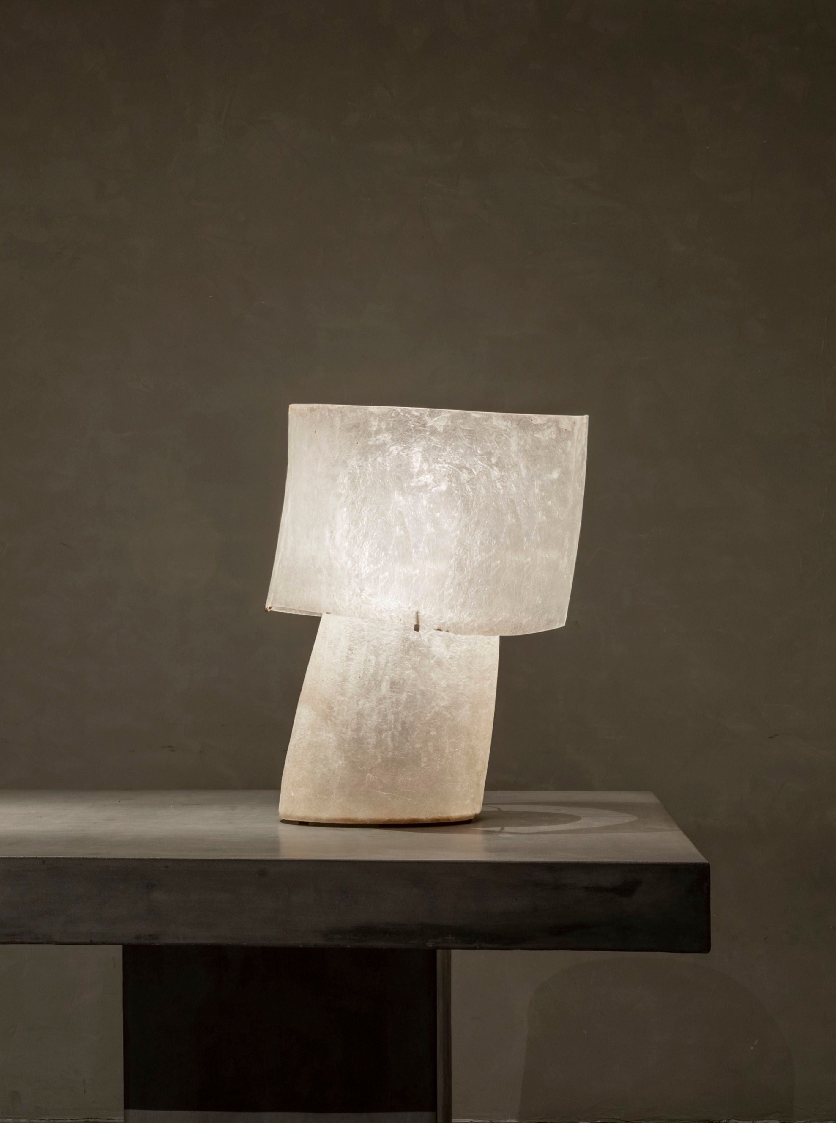 A uniquely designed mush lamp from Kilzi, known as "The Mush Table Lamps - Tall," with its translucent, tilted structure, rests elegantly on a dark minimalist table against a plain backdrop. Constructed from fibreglass, the lamp provides a gentle, ambient glow.