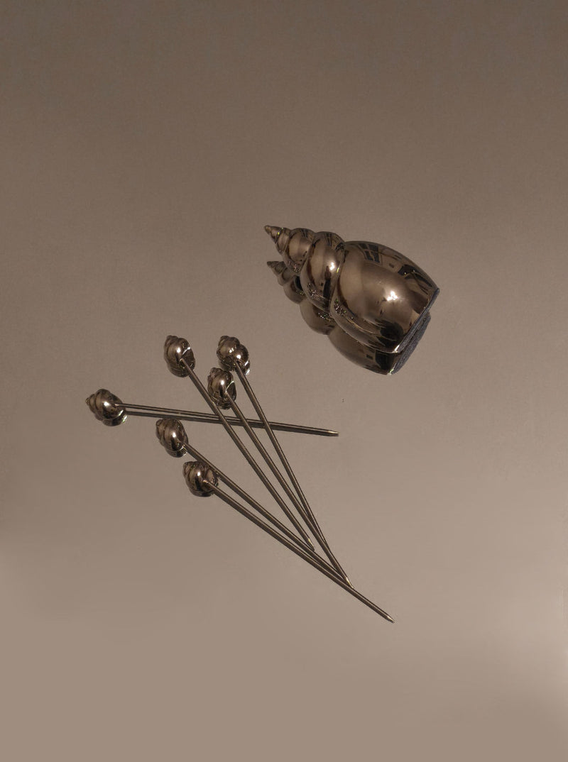 Les Objoies Shell Cocktail Picks feature a silver, conch-shaped shell holder beside six ocean-inspired picks with round decorative tips, displayed on a plain light brown surface.